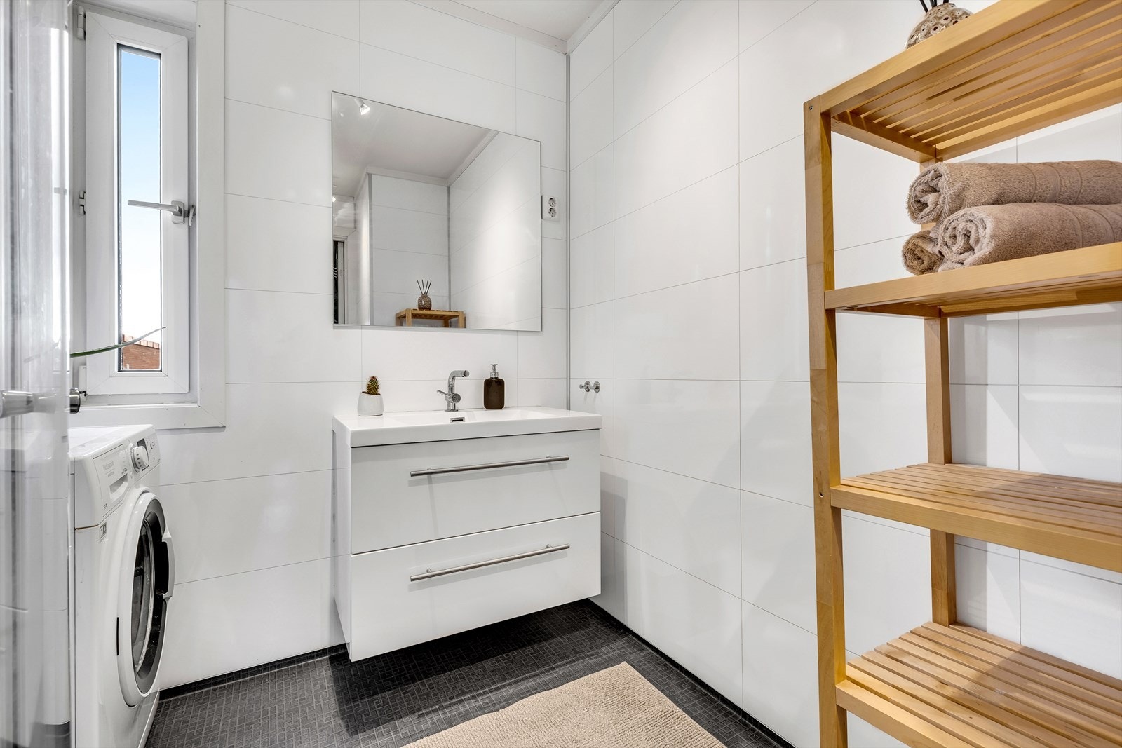 Baderomsinnredningen har godt med oppbevaringsplass for håndklær og baderomsartikler. Badet er ellers utstyrt med dusjhjørne, servant med underskap, opplegg for vaskemaskin, veggmontert WC og mekanisk avtrekk. Galleribilde