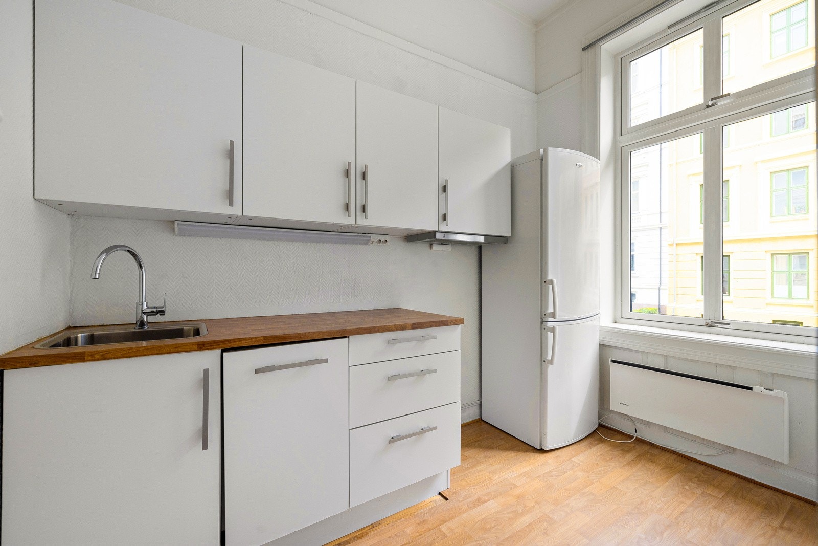 HTH-kjøkkeninnredning med slete hvite fronter, 28mm tykk laminatbenkeplate med imitasjon av finér og nedfelt vaskekum i børstet stål og ettgreps blandebatteri i rustfritt stål. Galleribilde