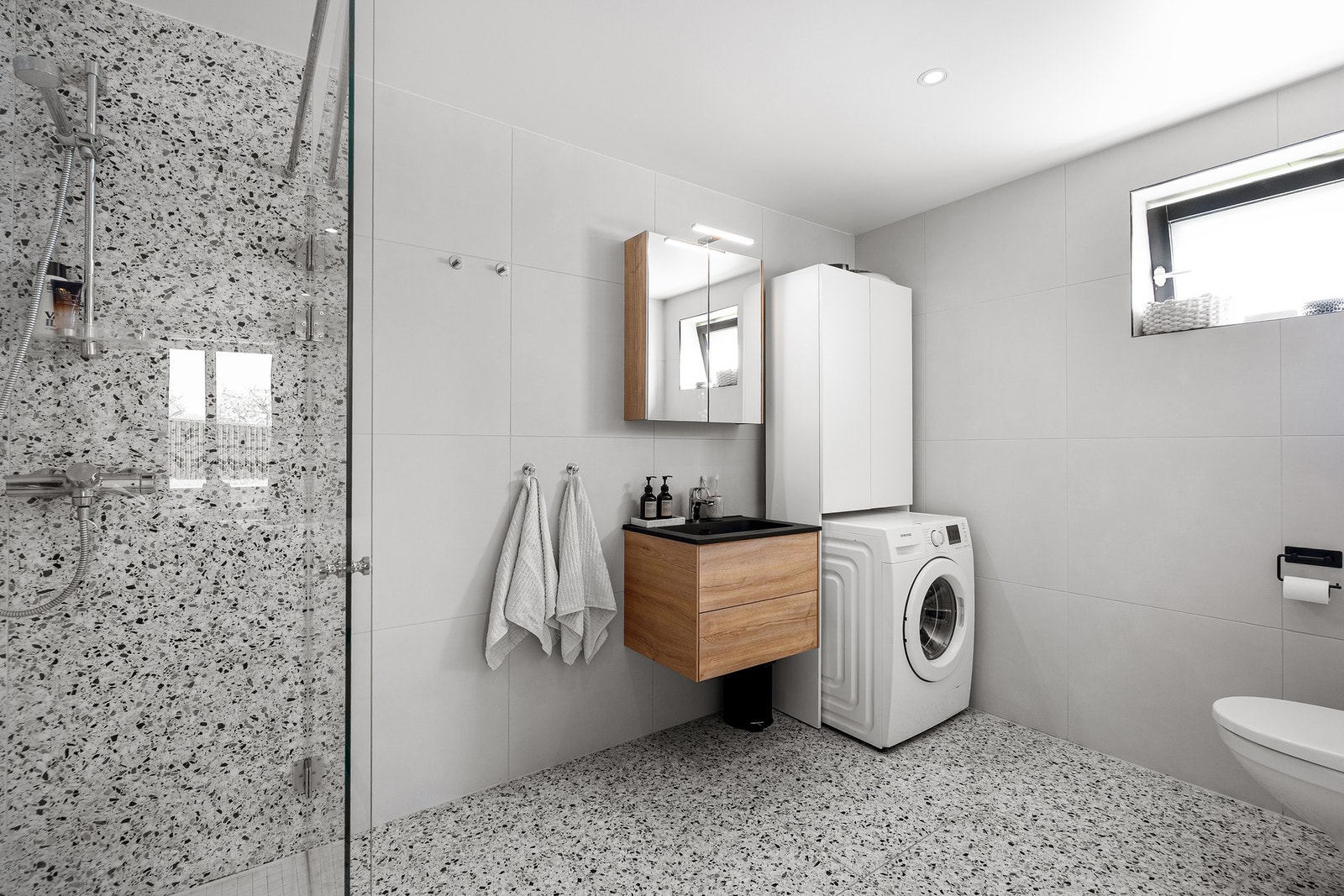 Flislagt bad med dusjhjørne, vegghengt toalett og opplegg for vaskemaskin Galleribilde