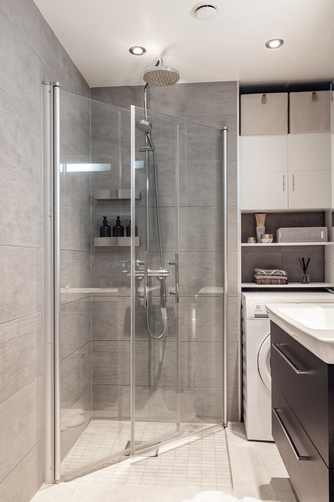 Badet er utstyrt med veggmontert toalett, dusjhjørne med innfellbare glassdører og bred baderomsinnredning med servant, ettgreps blandebatteri, skuffer og vegghengt speilskap. Galleribilde