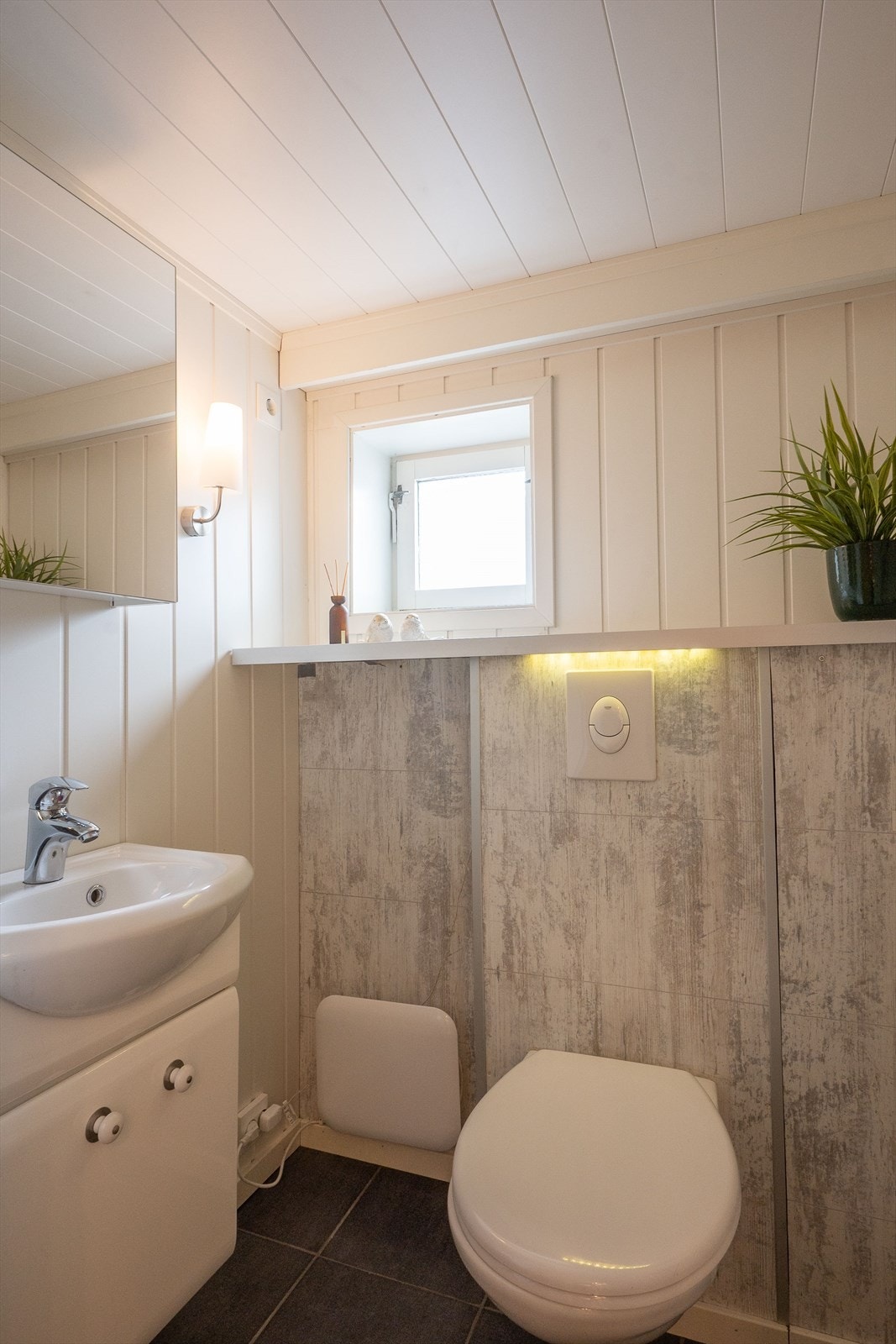 Vegg-i-vegg med badet ligger toalettrommet med flislagt gulv, varmekabler, toalett, servantskap og speilskap. En hylle gir plass til pynt eller oppbevaring. Galleribilde