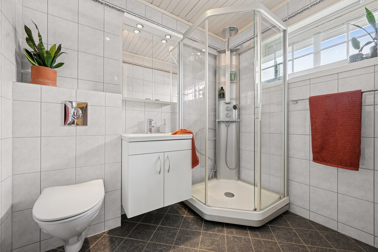 Pent bad med fliser på vegger og gulv med gulvvarme. Badet har baderomsinnredning med heldekkende servant og speil på vegg, vegghengt toalett og dusjkabinett. Galleribilde