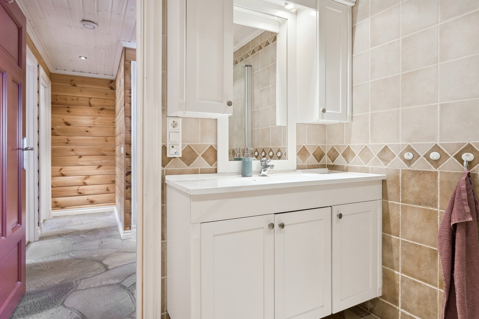 Lys baderomsinnredning med heldekkende servant, speil med belysning og vegghengte skap over. Baderommet er også utstyrt med dusjhjørne med innfellbare glassdører og vegghengt toalett. Galleribilde