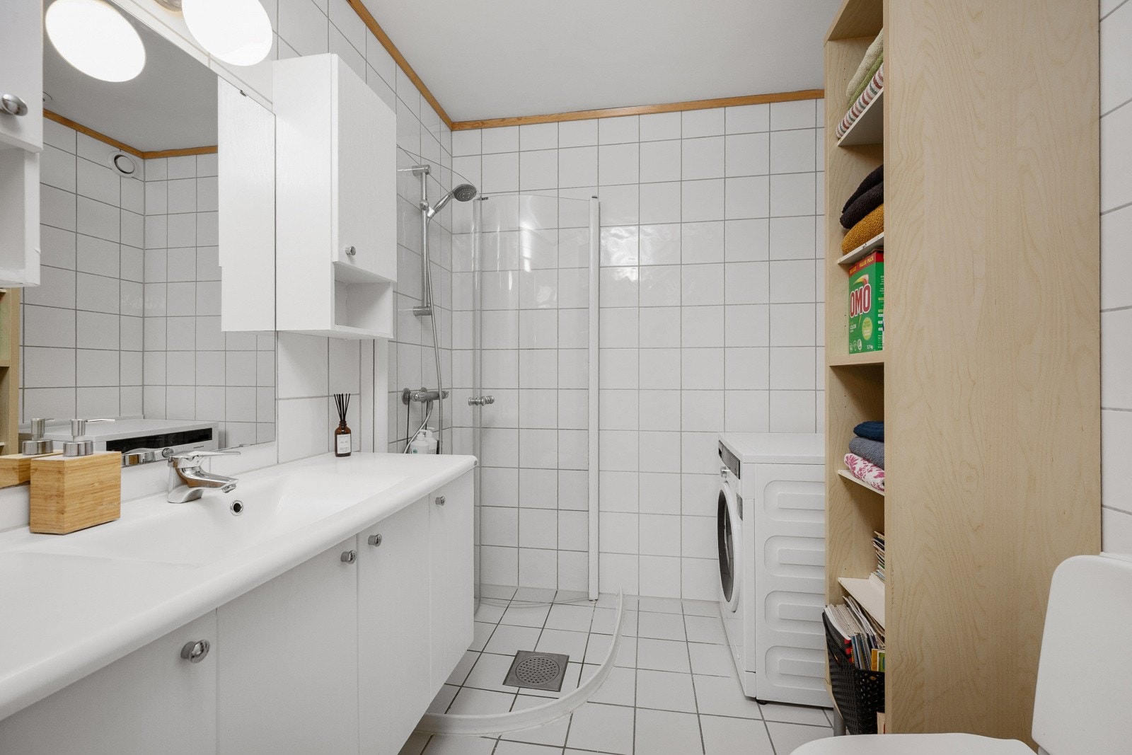Baderommet har fliser på vegger og gulv med gulvvarme. Badet har en innredning med heldekkende servant med underskap, speil med lys, og overskap til høyre for speilet. I tillegg har badet dusjhjørne med glassdører, toalett og opplegg for vaskemaskin. Galleribilde
