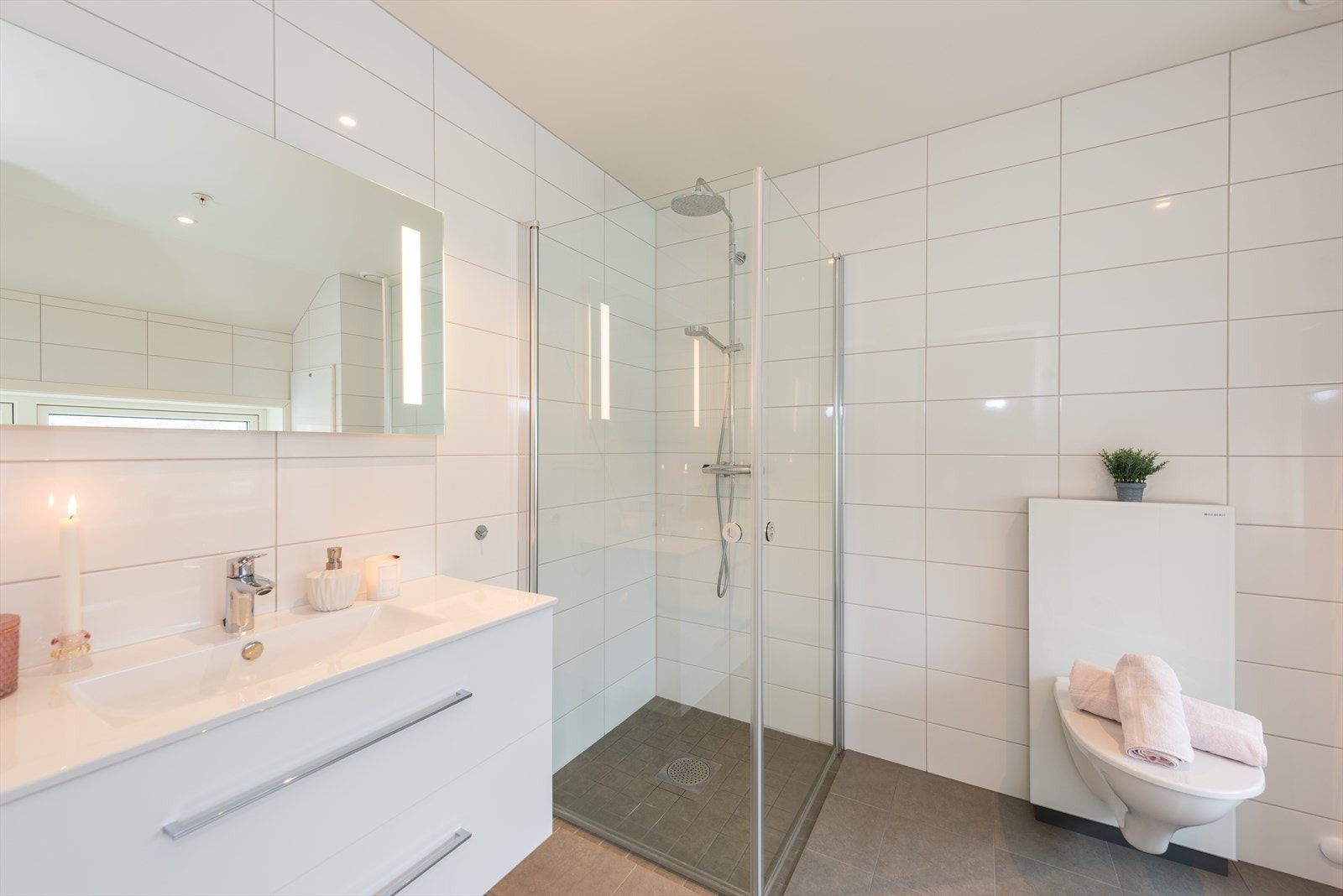 Badet har flislagt gulv og vegger. Rommet er utstyrt med vegg hengttoalett, enkelt servant og dusj med innfellbare dører. Galleribilde