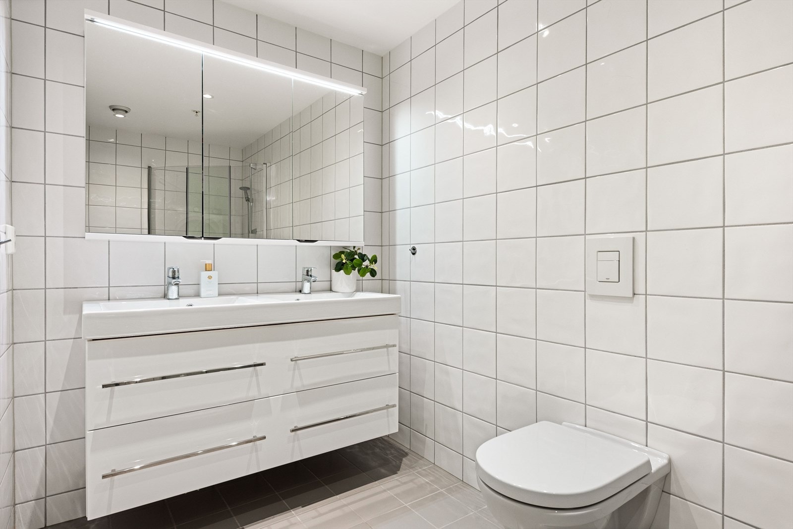 Videre kommer man inn i et lyst og flislagt bad som er har innredning med dobbel servant, veggskap med speil og integrert belysning, toalett og dusjhjørne. Galleribilde