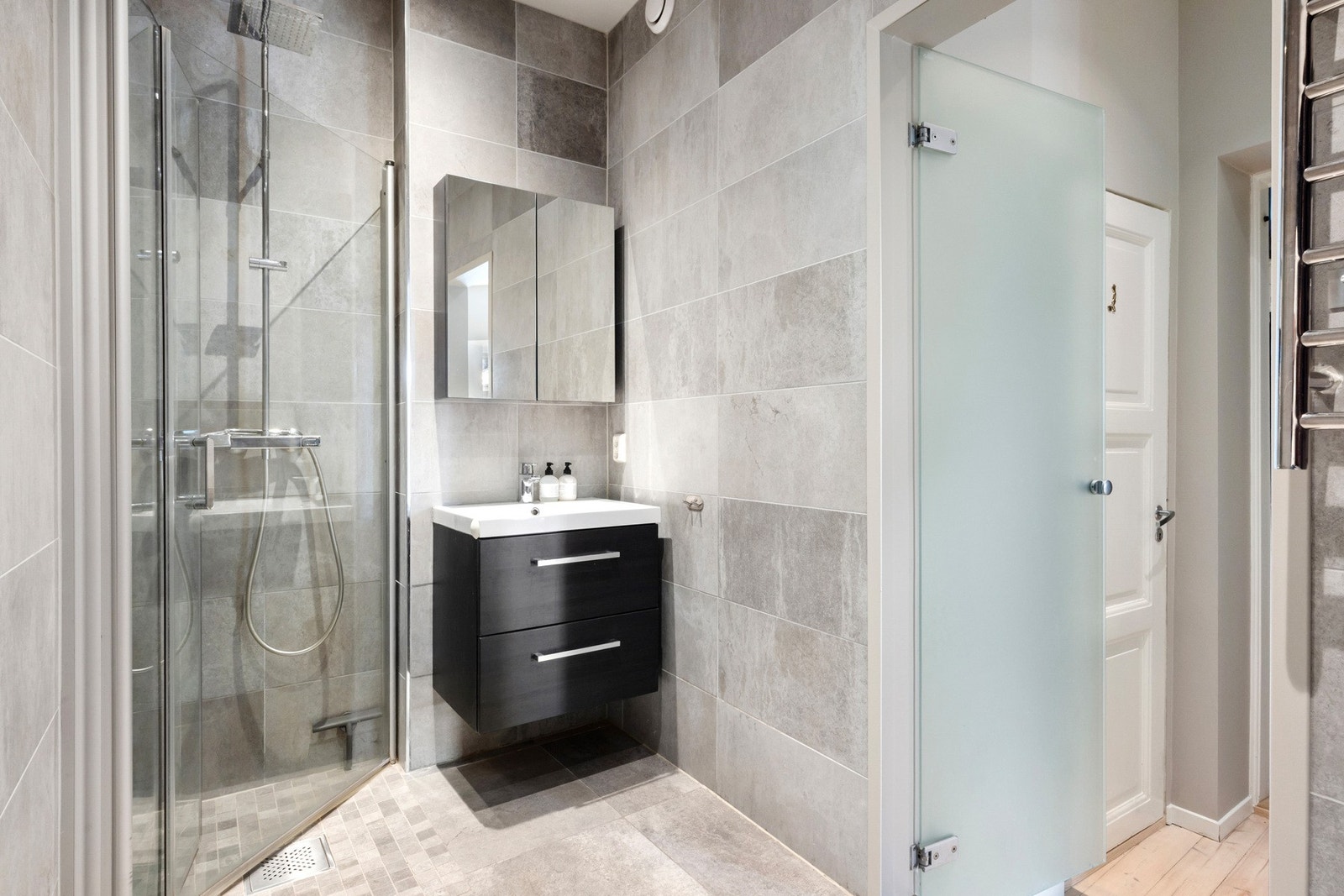 Pent og stilrent bad med flotte fliser i gråtoner. Badet er helfliset og har en romslig dusjsone med glassdører. Praktisk servantskap med oppbevaringsplass og speilskap med integrert belysning. Varmekabler i gulvet gir ekstra komfort. Et funksjonelt og de Galleribilde