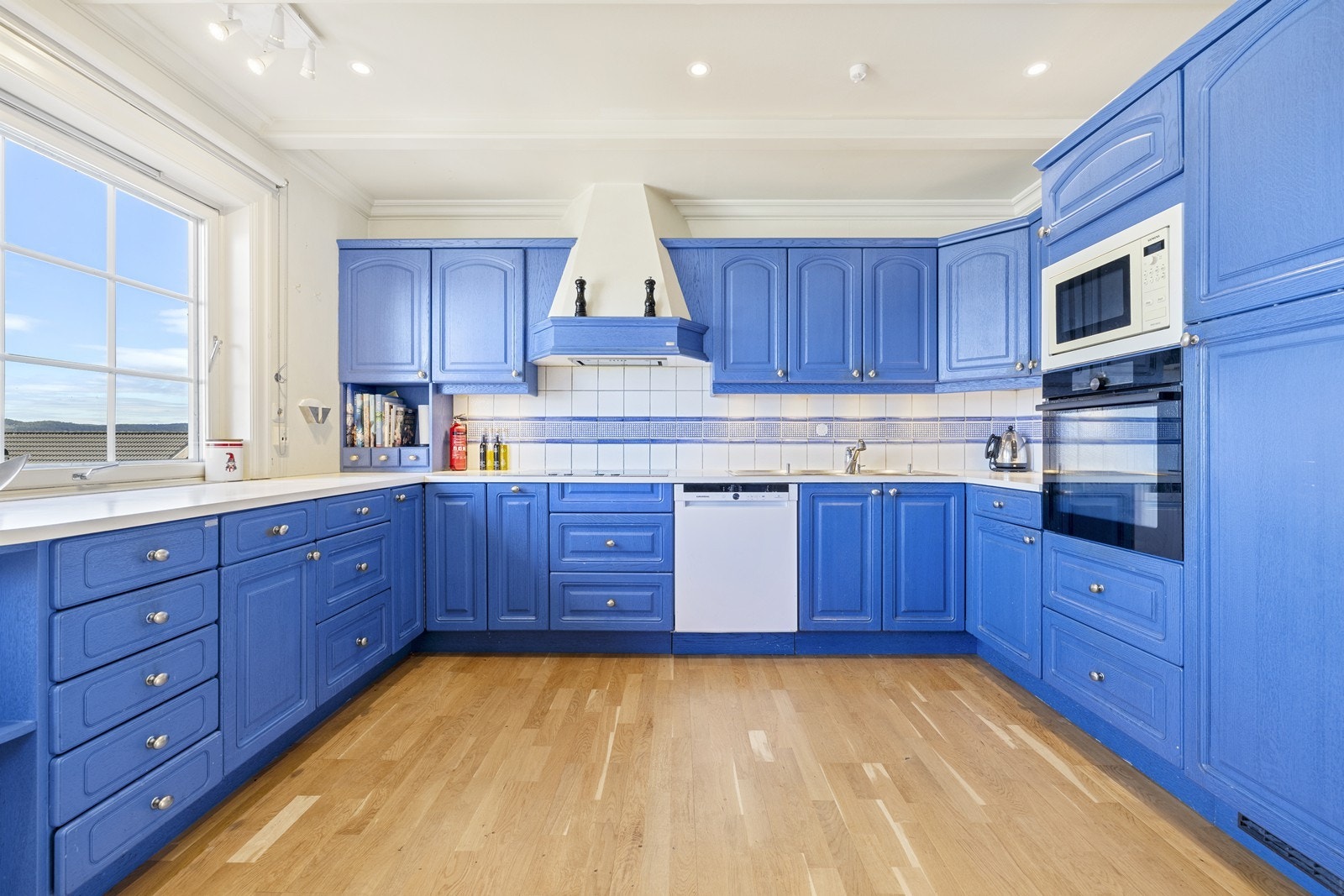 Stort kjøkken malt i blått med en lys benkeplate av laminat og flisfelt over. Innredningen er profilert, og byr på rikelig med benke- og skapplass. Galleribilde
