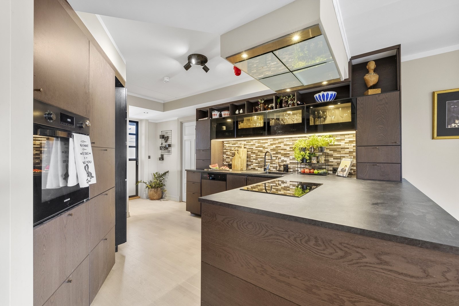 Det er mørke fronter i trestruktur og en komplimenterende mørk benkeplate. Over benkeplaten bak vasken er det lagt kule fliser. Galleribilde