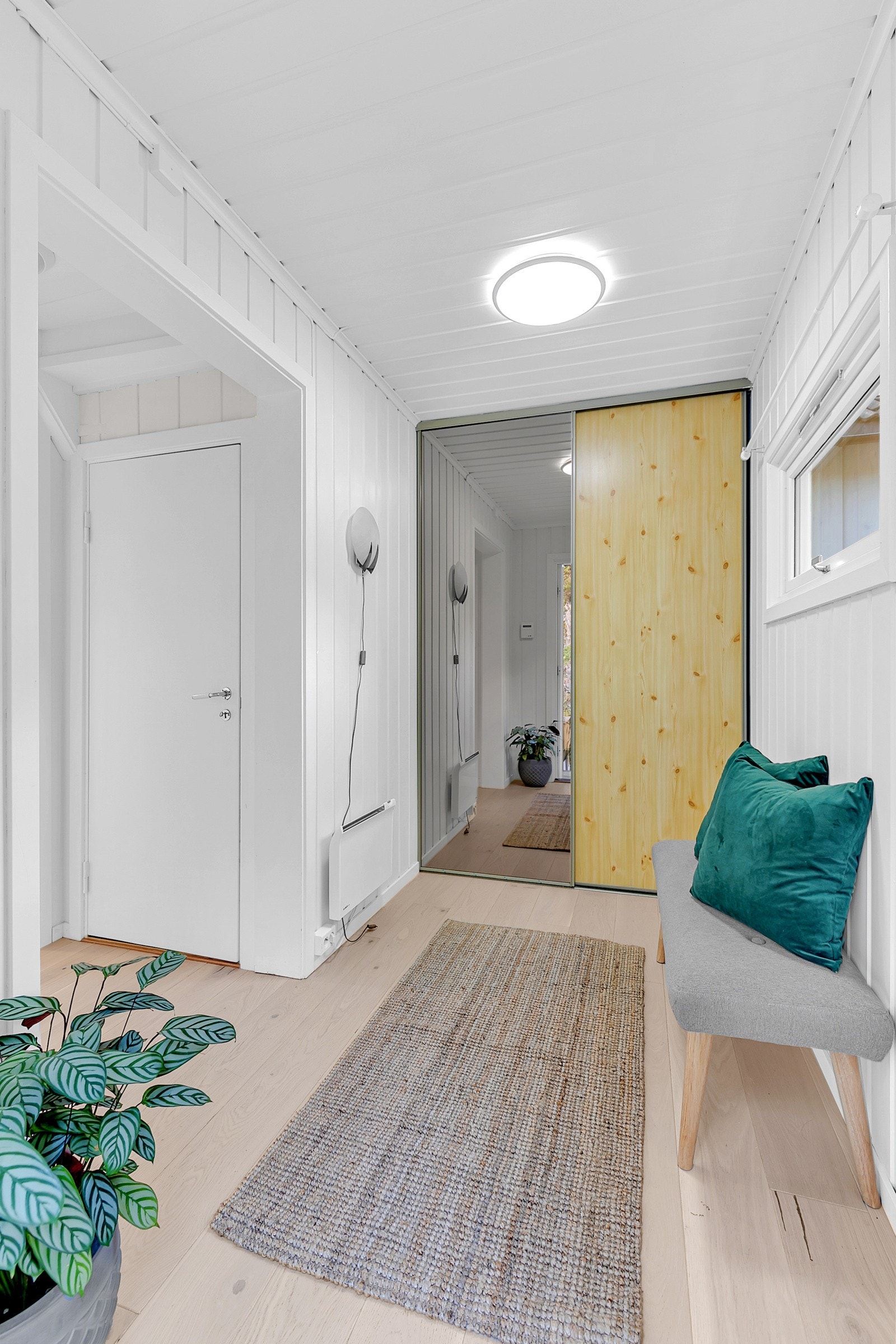 Praktisk inngangsparti med skyvedørsgarderobe. Vegger og himling med hvitmalt panel, enstavsparkett på gulv. Stor åpning mot mellomgang og vindu som gir gjennomgående lys inn mot stuen. Galleribilde