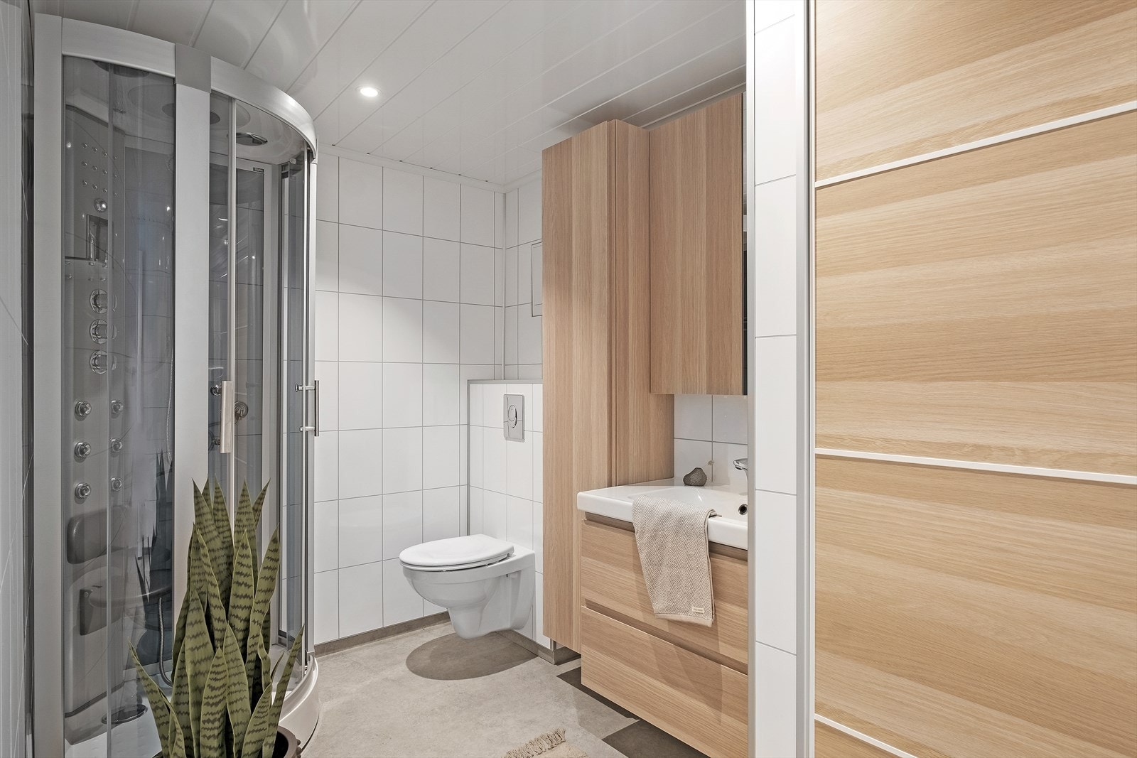Badet har vinylbelegg med varmekabler på golv, baderomsplater på veggar og panel med downlights i himling. Galleribilde