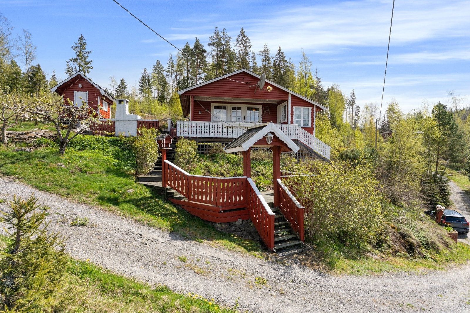 Stor eiet tomt. Rødt hytte med hvite detaljer, veranda og trapp. Grusvei foran og omgitt av vegetasjon. Galleribilde