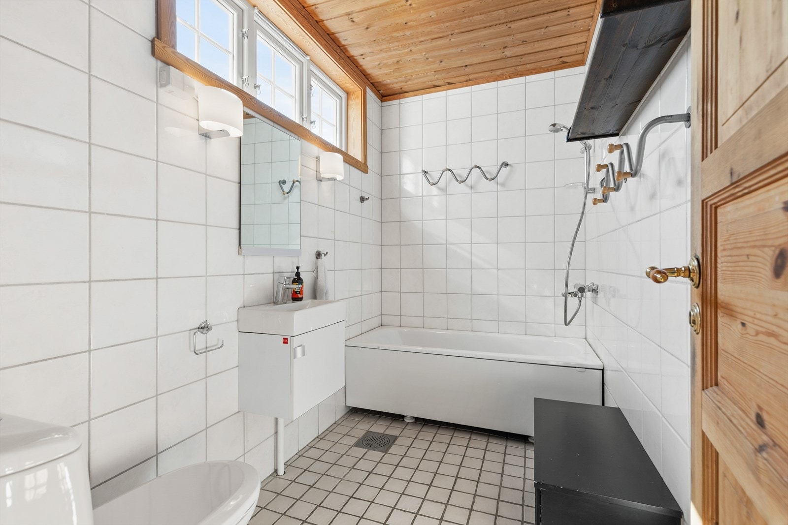 Baderom med flislagte gulv og vegger. Badet består av toalett, servant med skap og badekar med dusjløsning. Galleribilde