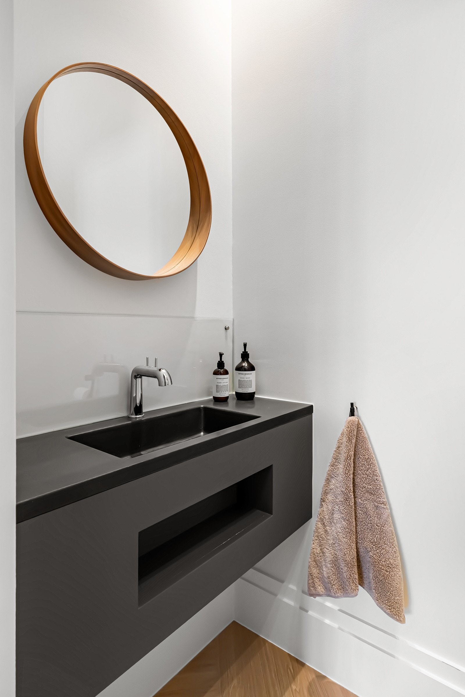 Servantinnredningen er mørk med matt front, heldekkende servant, rundt speil og plate i pleksiglass mot vegg. Galleribilde