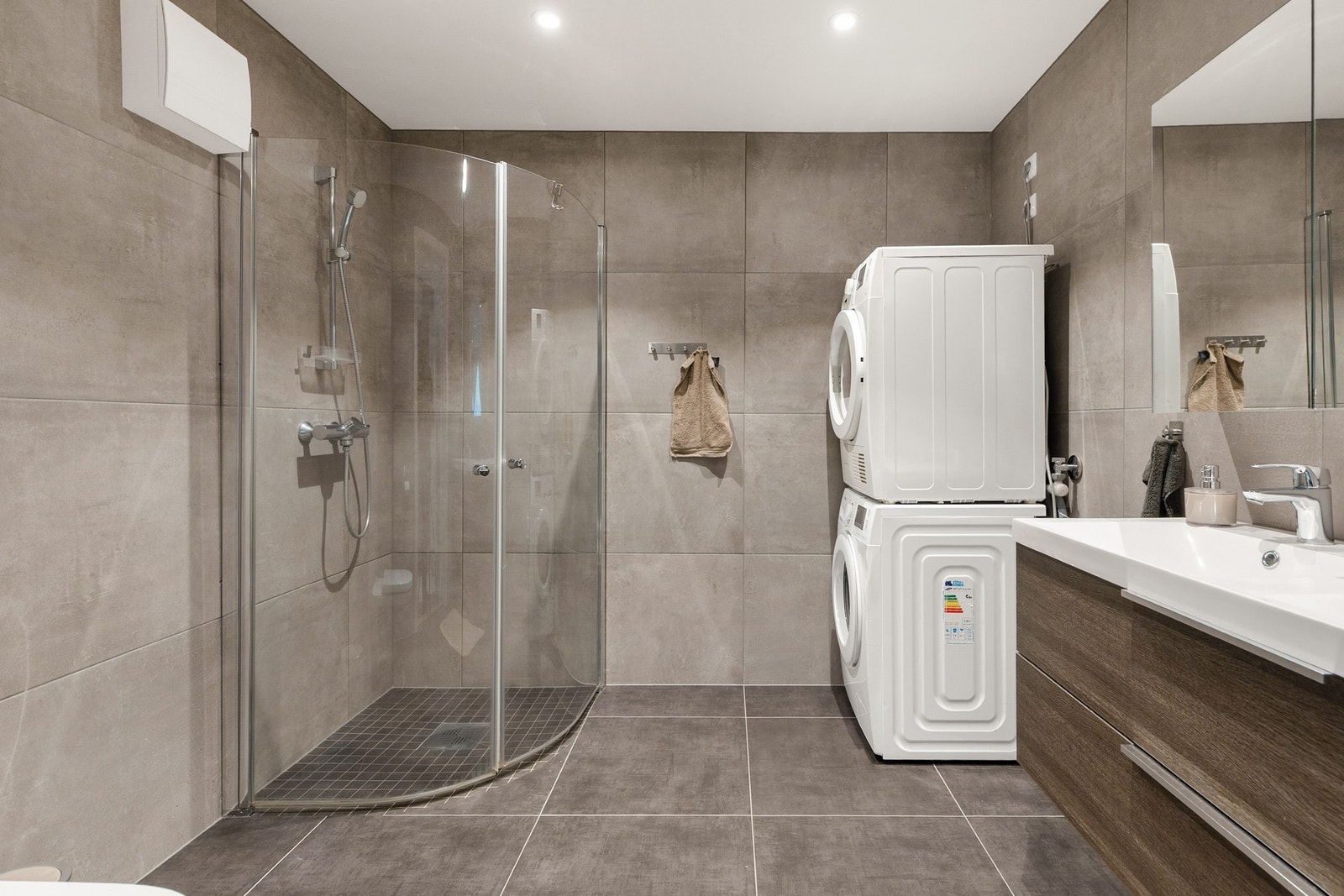 Badet har et dusjhjørne med glassdører og vegghengt dusjarmatur, opplegg til vaskemaskin samt et vegghengt toalett Galleribilde