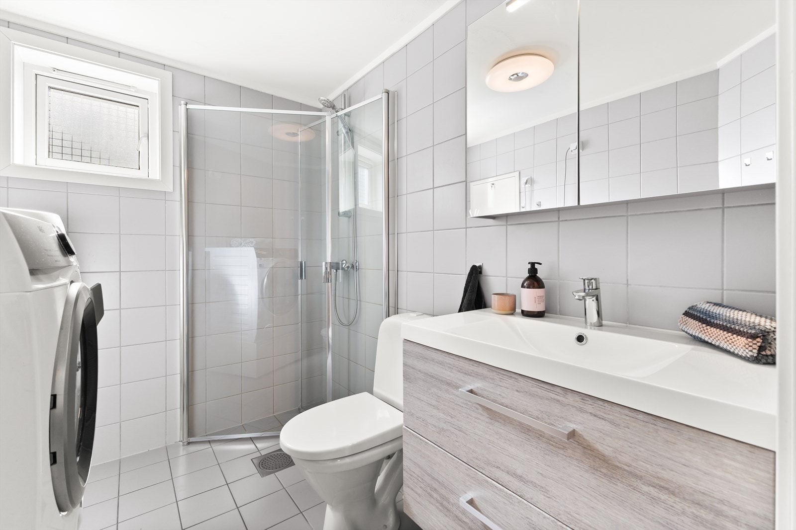 Badet har innfellbare dusjdører, toalett, servant og speilskap med innredning og opplegg for vaskemaskin Galleribilde