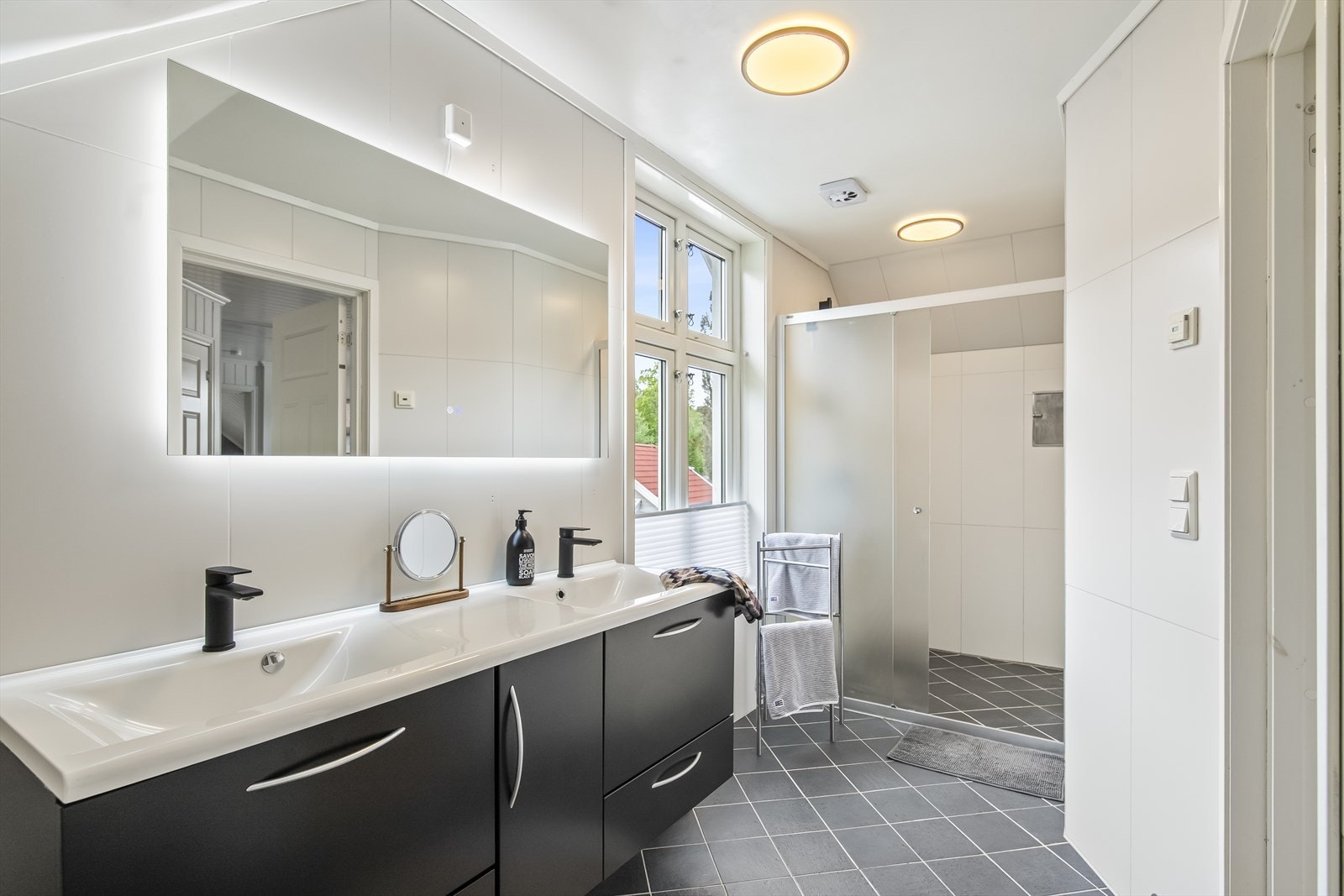 Her finner du både et komfortabelt badekar med dusjløsning og en romslig dusjnisje med skyvedør. Innredningen er stilfull med dobbel servant, skuffeløsninger i grått, veggskap og speil med integrert lys. Galleribilde