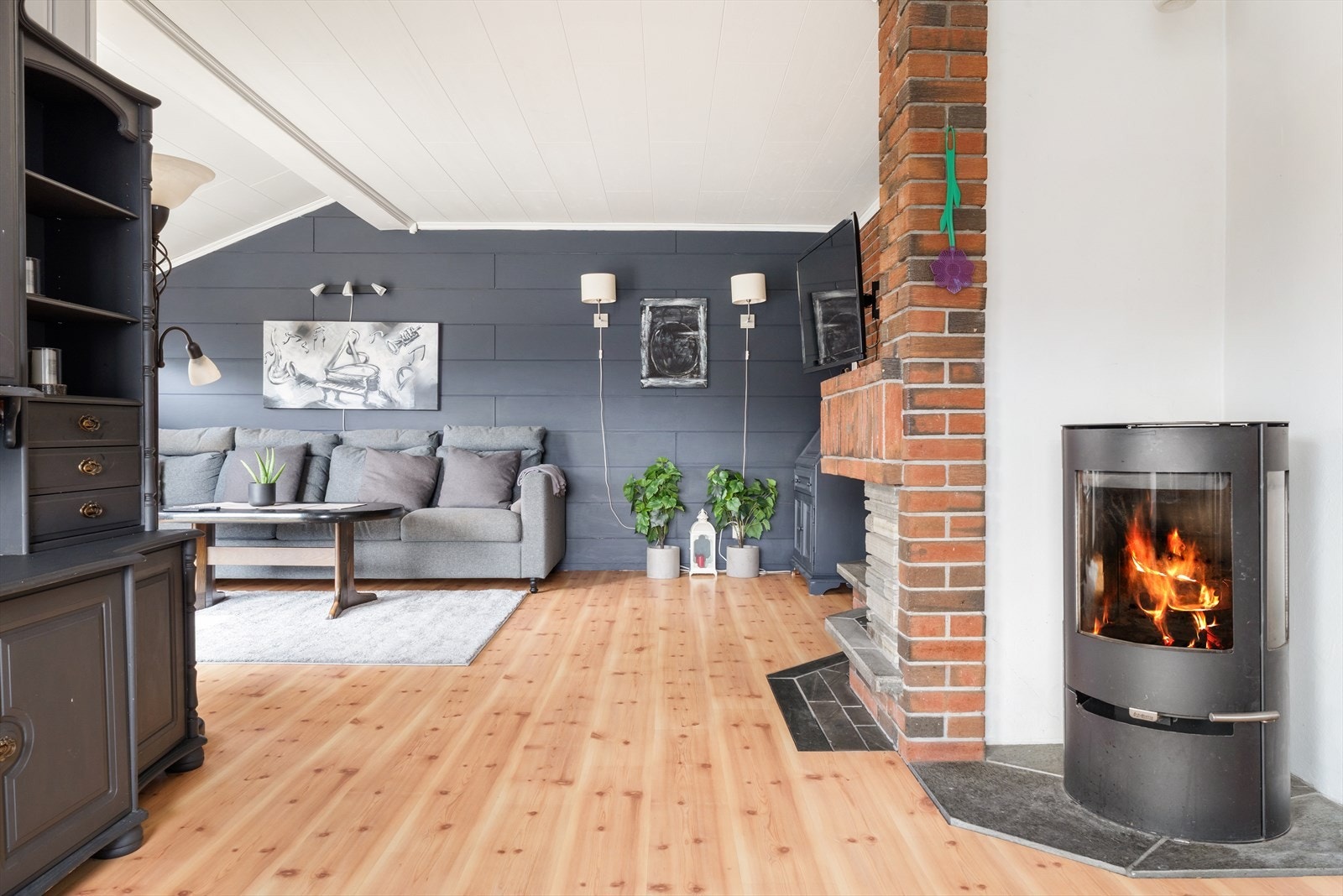 Romslig stue med laminat på gulv og malt panel og malte plater på vegger. Det er både vedovn og peis i stuen med stor teglsteinsmur Galleribilde