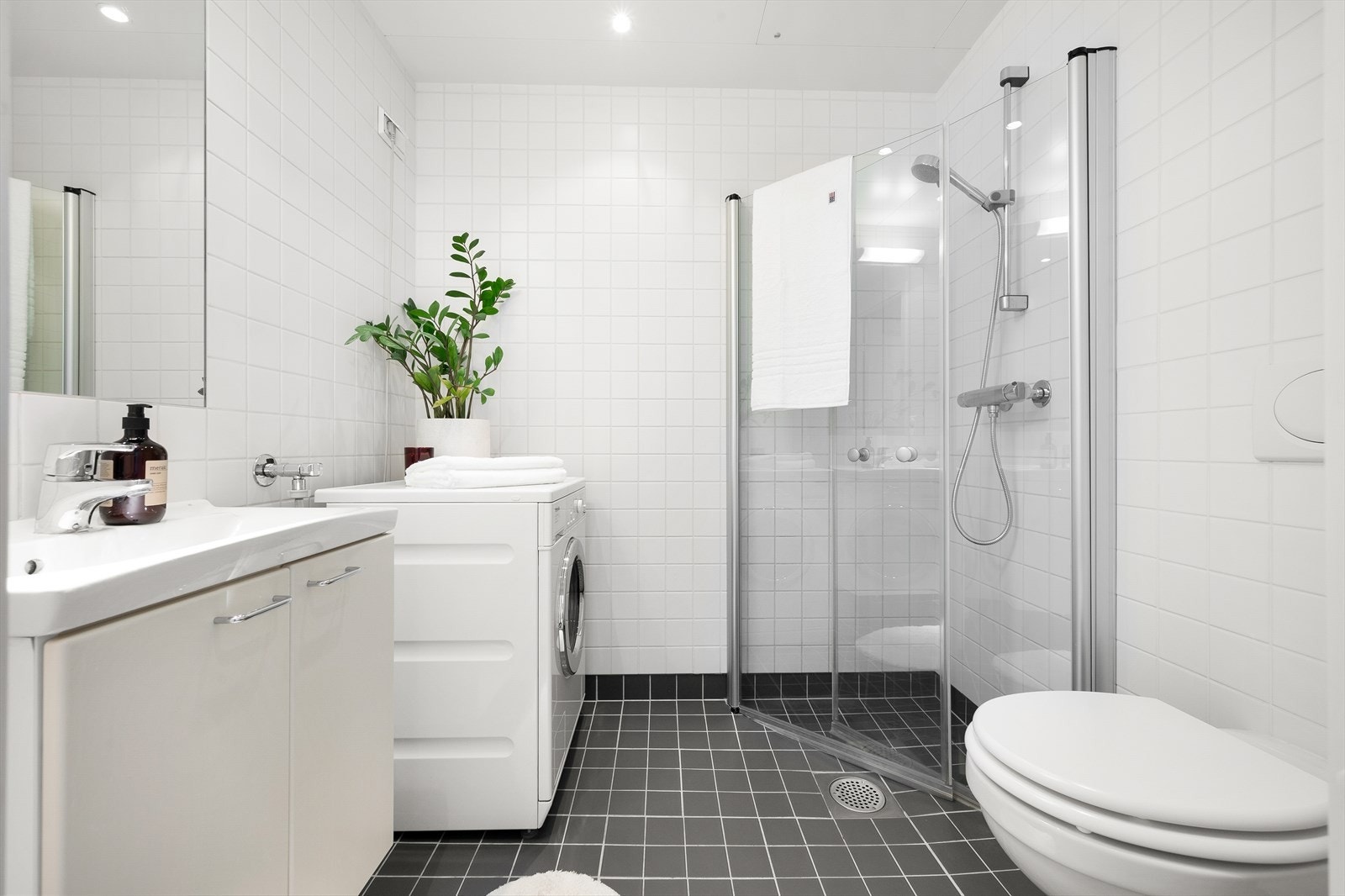 Romslig og tidløst bad med fliser, gulvvarme og opplegg for vaskemaskin. Rommet er innredet med veggmontert toalett, dusjhjørne med innfellbare glassdører og baderomsinnredning med skap, servant og speil. Galleribilde