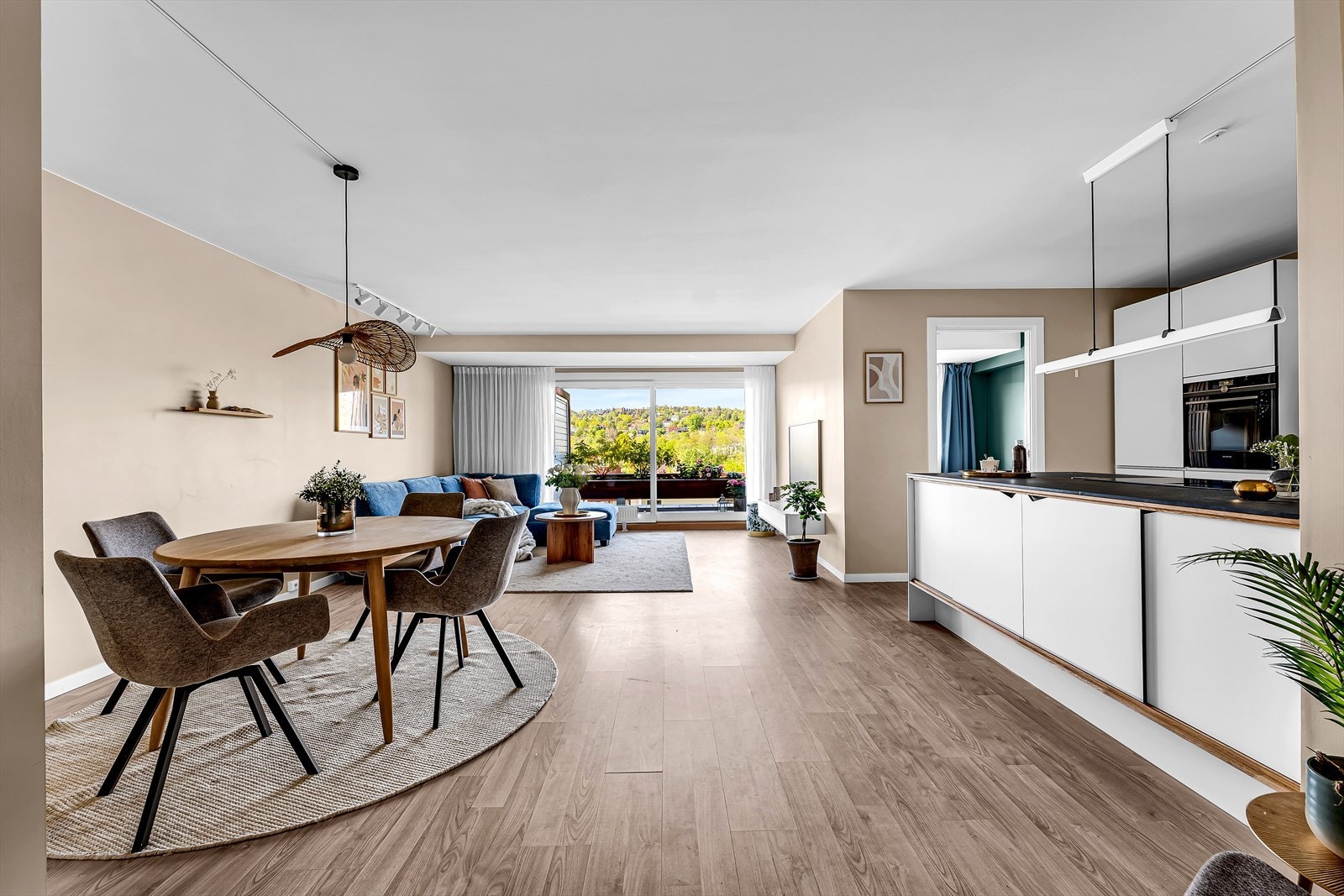 Den åpne kjøkkenløsningen mot spisestuen skaper en luftig og innbydende atmosfære, hvor naturlig lys strømmer inn gjennom store vindusflater og gir en harmonisk forbindelse til de grønne omgivelsene utenfor. Galleribilde