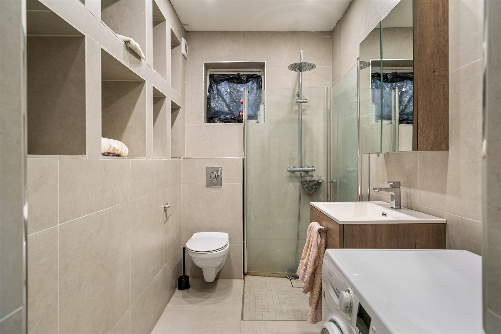 Flislagt bad fra 2012 med veggmontert toalett, dusjhjørne med nedsenk og innsvingbare glassdører. Badet har opplegg for vaskemaskin og nisjer i veggen. Formstøpt servant med laminat underskap og skuffer, samt speilskap. Galleribilde