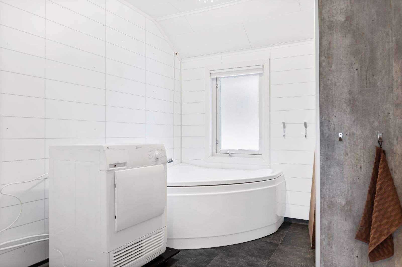 Bad med plass til stort badekar, dusnisje like bak veggen til høyre og plass til både vaskemaskin og tørketrommel. Galleribilde