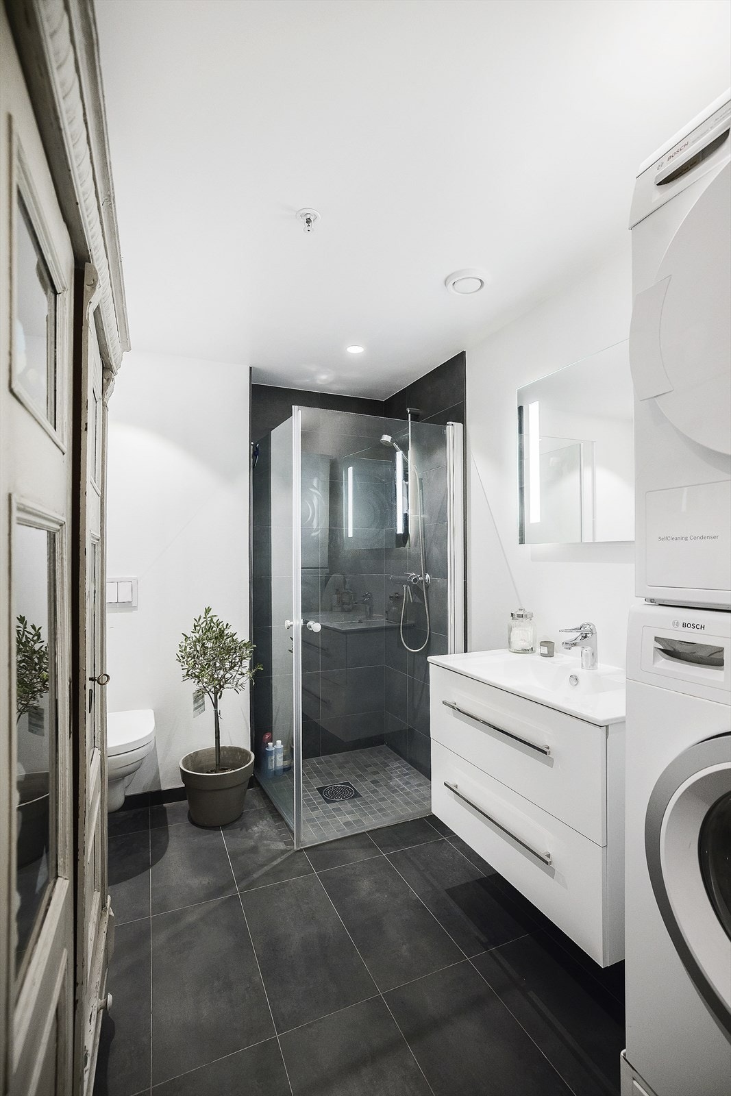 Baderommet er utstyrt med heldekkende servant over skuffeinnredning, vegghengt toalett, innfellbare dusjdører som gir et dusjhjørne, samt opplegg til vaskemaskin og tørketrommel Galleribilde