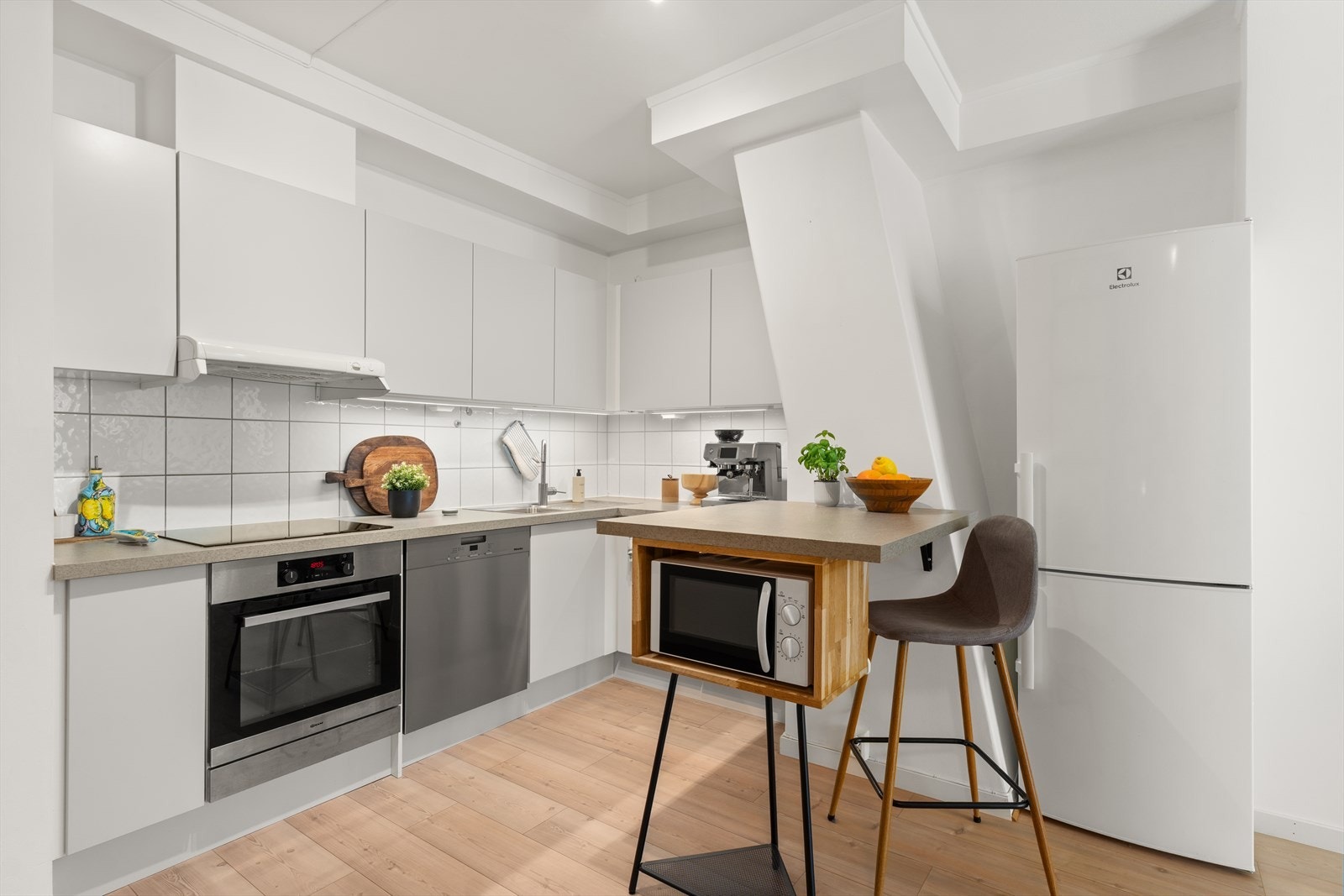 Kjøkkenet har en moderne og funksjonell utforming med en L-formet innredning som gir god arbeidsflyt og lagringsplass. Innredningen består av hvite, glatte skapfronter med håndtaksløse løsninger for et stilrent uttrykk Galleribilde