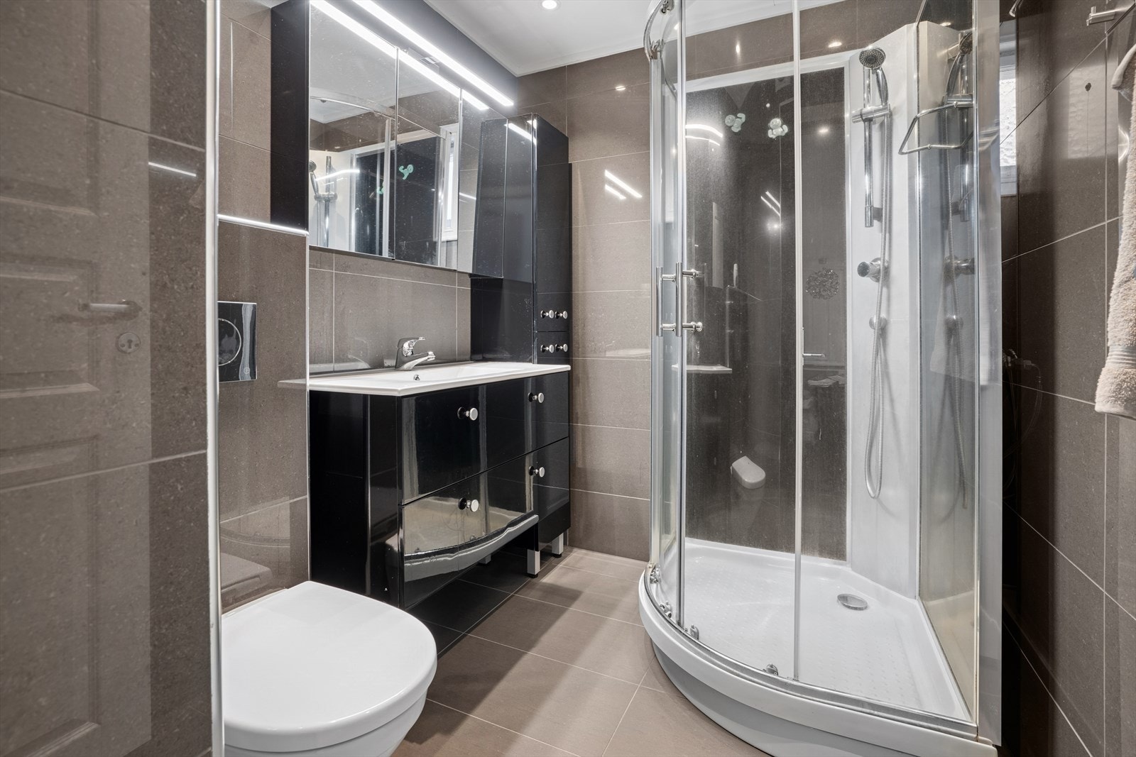 Bad med flislagte vegger og gulv. Vegghengt klosett, dusjkabinett og servant med innredning. Galleribilde