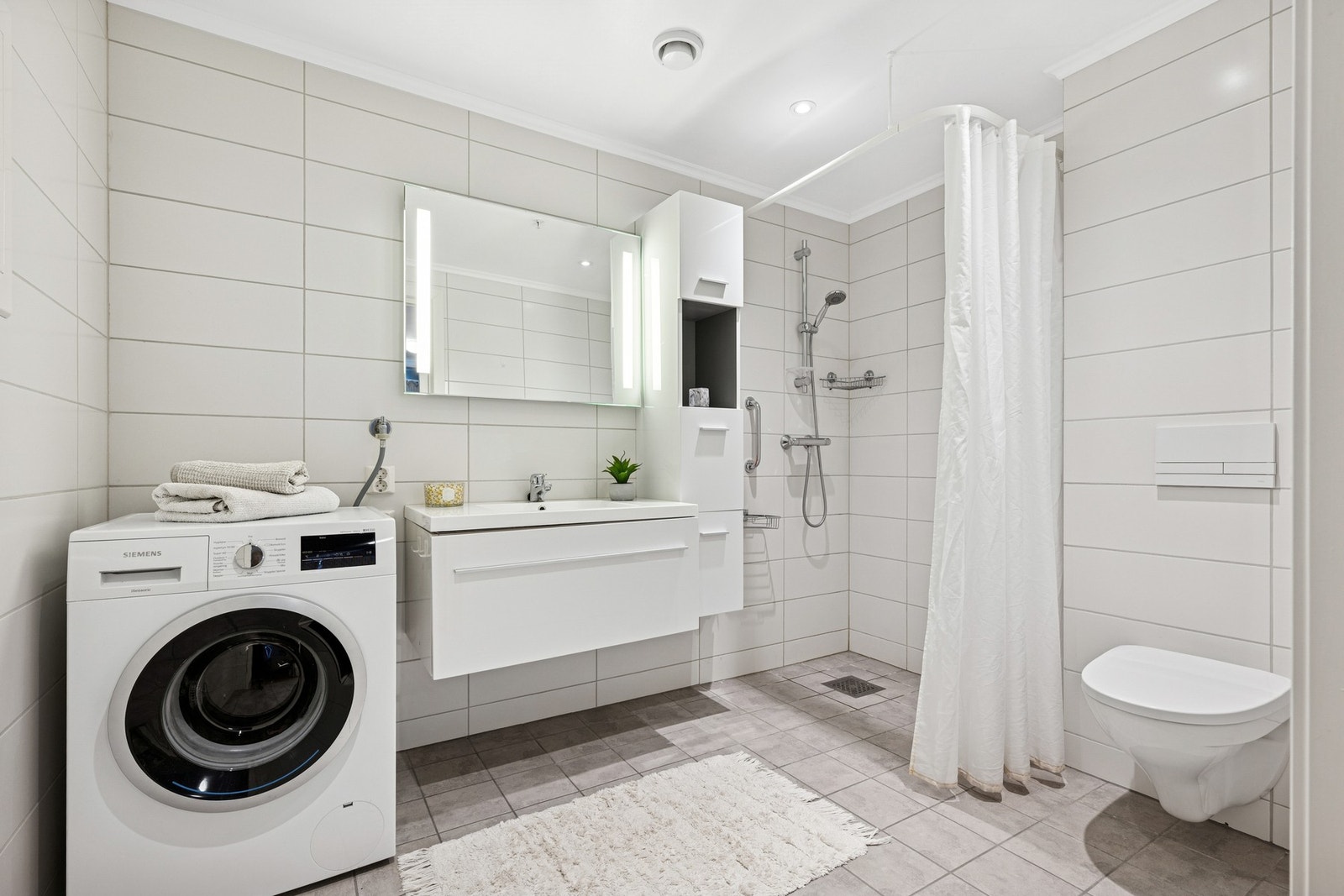 Lyst og romslig baderom med dusjhjørne, vegghengt toalett, servant og opplegg for vaskemaskin og tørketrommel. Galleribilde