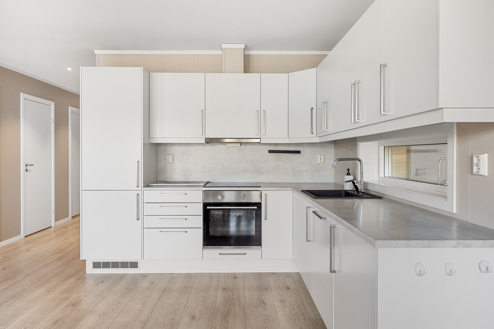 Innredningen har hvite glatte fronter og lys laminert benkeplate med nedfelt antrasitt oppvaskkum. Praktisk med plater i benkeryggen ved platetoppen og belysning under overskapene. Galleribilde