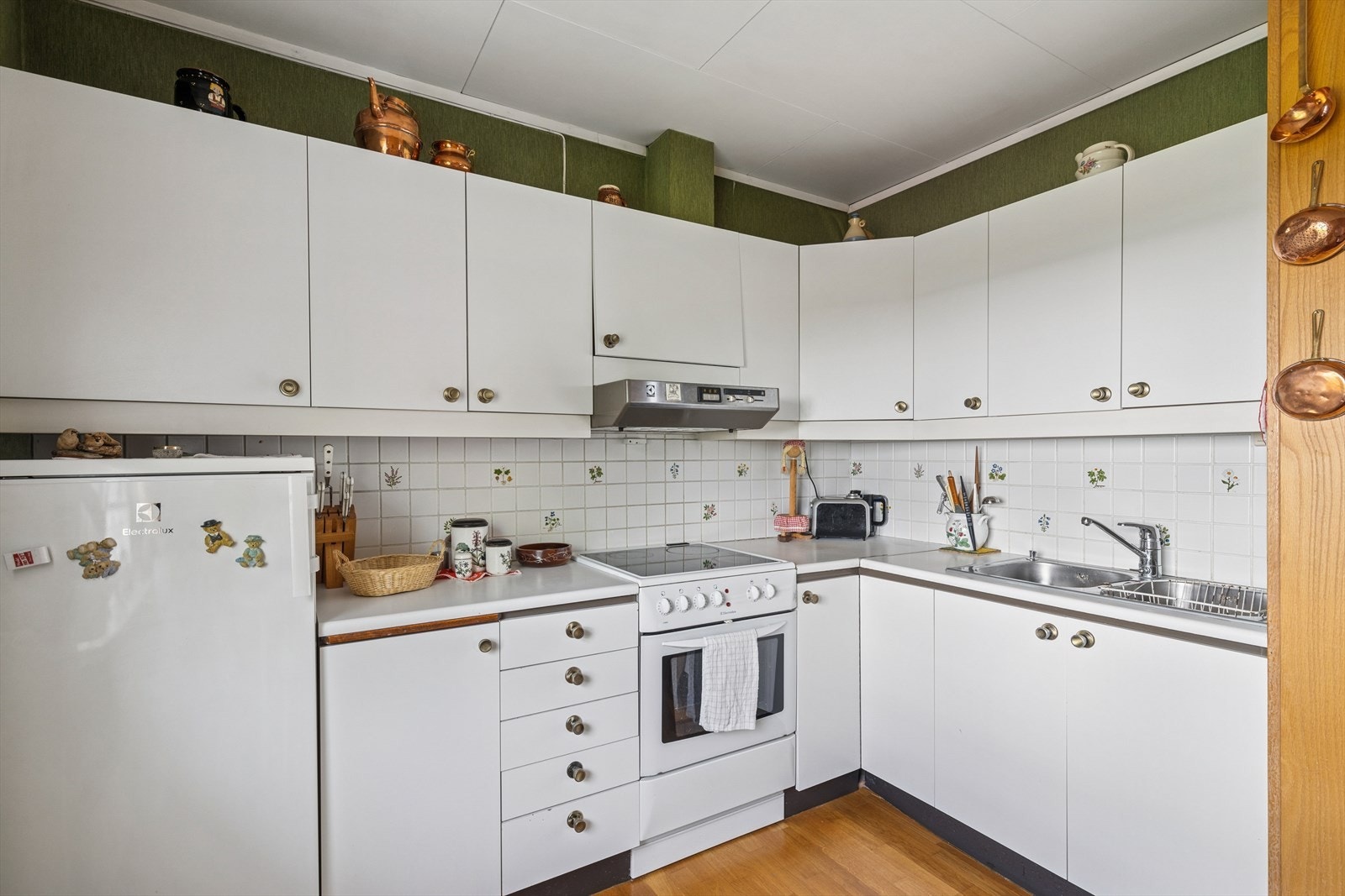 Kjøkkeninnredning med glatte folierte fronter, laminat benkeplate med nedfelt kum i rustfritt stål, fliser på vegg over benk og ventilator tilsluttet kanal. Galleribilde