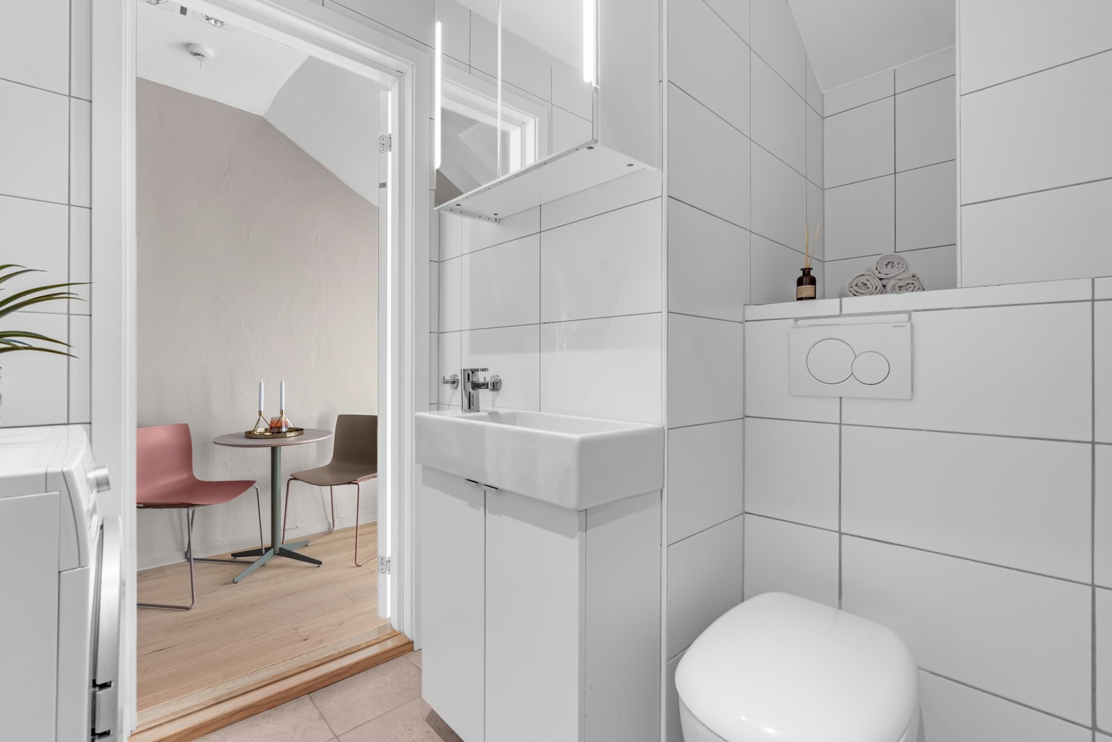 Vegghengt servantskap med dører. Ovenpåliggende servant med ett-greps armatur. Speilskap med belysning på vegg over servant, dusjhjørne med veggmontert hånddusj og vegghengt toalett med innebygget sisterne. Galleribilde