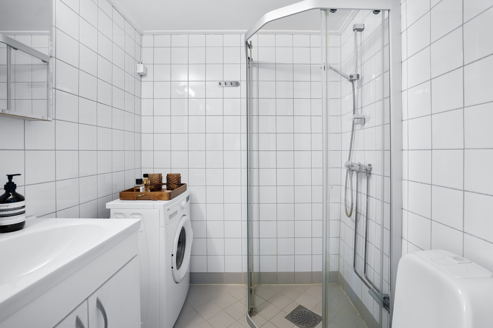 Flislagt baderom med opplegg for vaskemaskin, gulvvarme og malt himlingsflate. Badet er utstyrt med vegghengt servantinnredning med dører og ovenpåliggende servant med ett-greps armatur, gulvstående toalett og speilskap med overlys på vegg over servant. Galleribilde