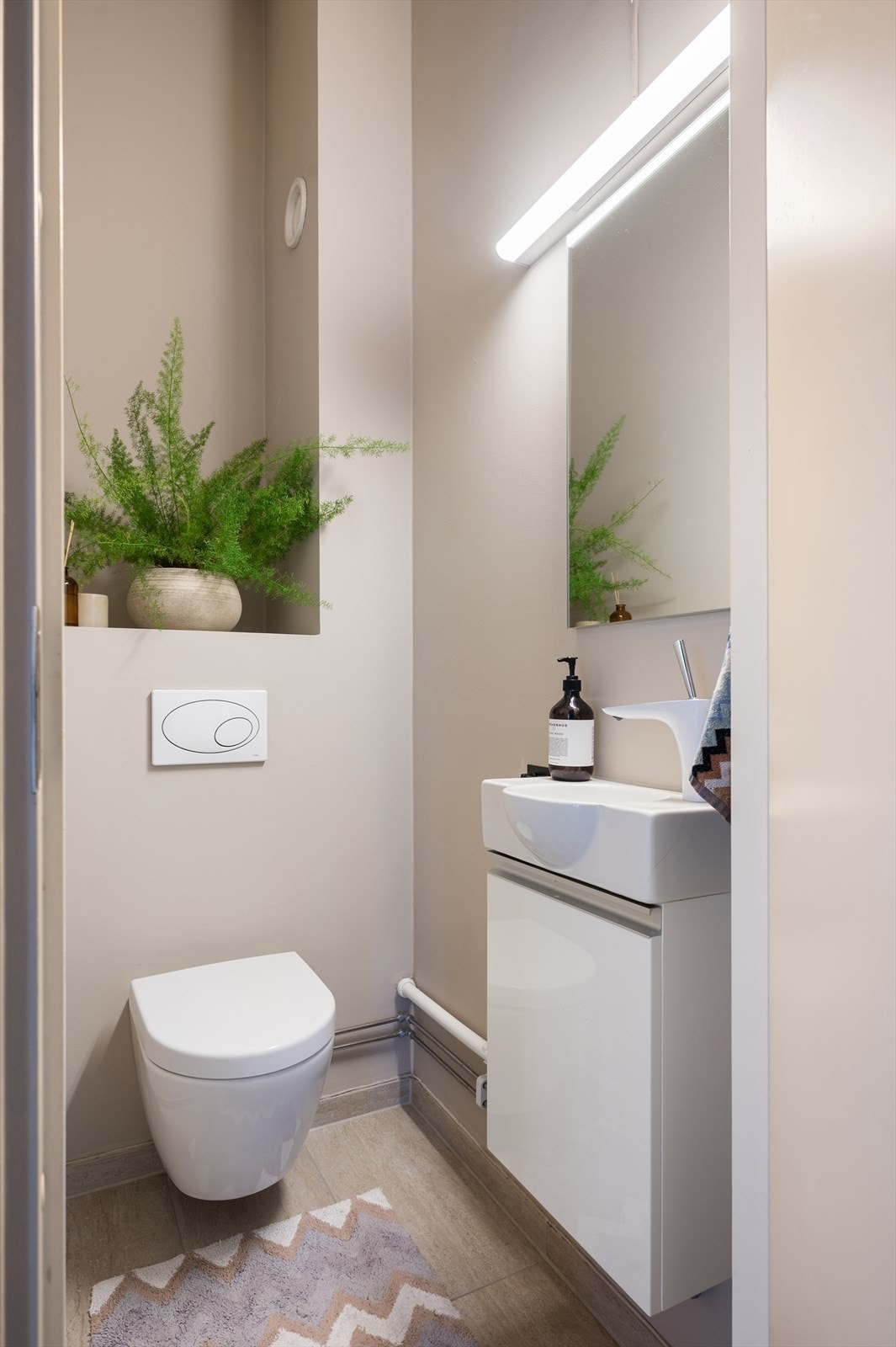 Praktisk toalettrom i forbindelse med entré. Gulvet er flislagt og innredningen består av vegghengt toalett og servant på innredning. Speil med belysning på vegg over servant. Rommet er utstyrt med mekanisk avtrekk. Galleribilde