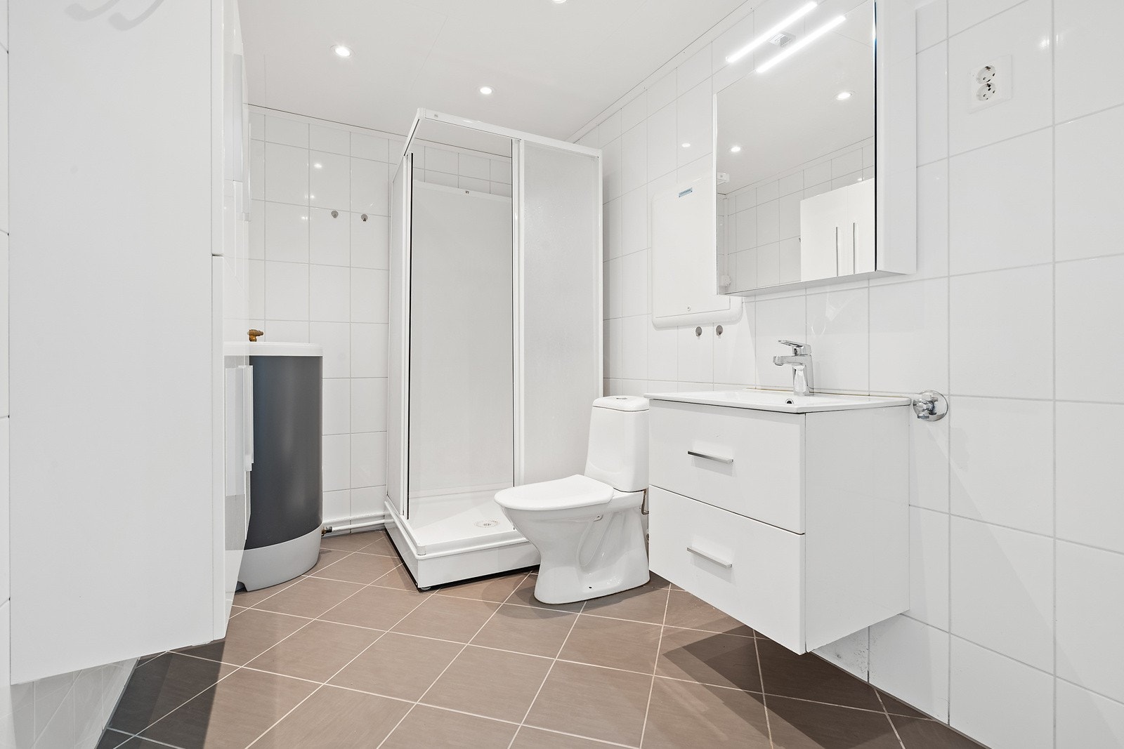 Badet fremstår tidløst med en lys utførelse og har blant annet dusjkabinett, toalett, servantskap og opplegg for vaskemaskin. Her får du også plass til en tørketrommel, og du finner leilighetens varmtvannstank i hjørnet. Galleribilde