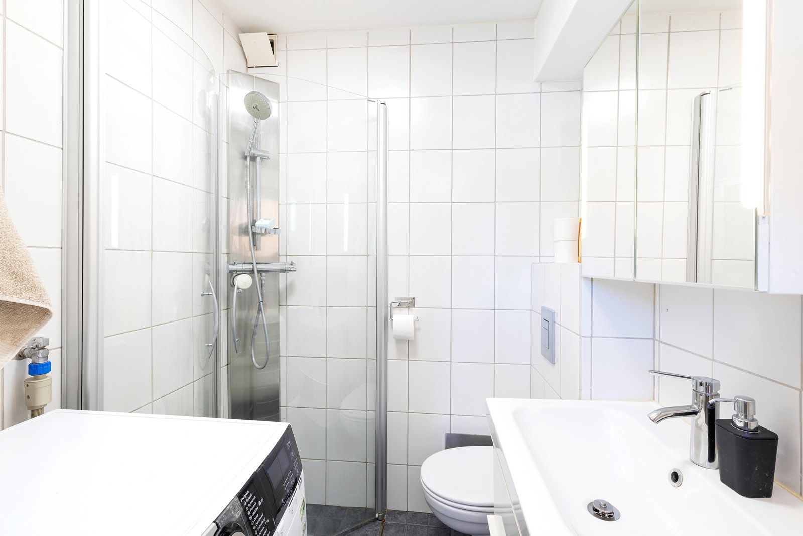 Flislagt bad med varme i gulv. Badet har dusjhjørne, veggehengt toalett, servant med underskuffer og opplegg til vaskemaskin. Galleribilde