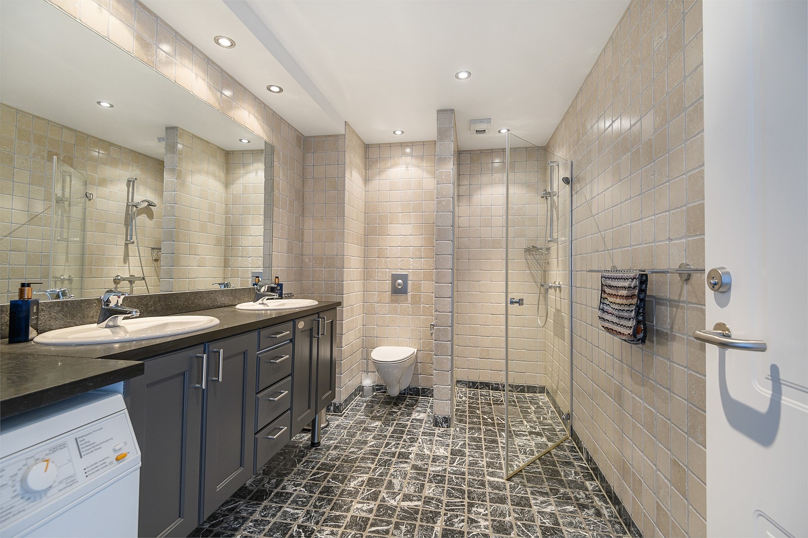 Badet har velholdte fliser i lyse og mørke farger, downlights i tak og varme i gulvet. Innredningen er romslig og består av dobbelt servantskap og et innfliset speil. Galleribilde