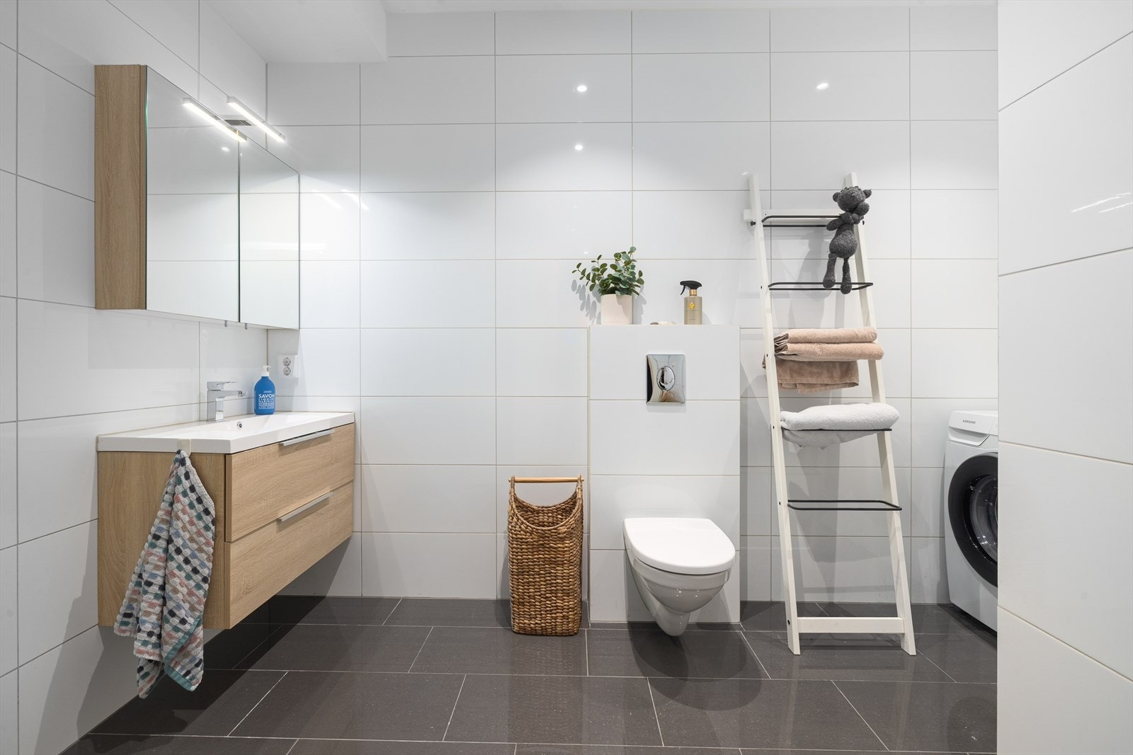 Badet inneholder dusjhjørne med innfellbare glassdører og veggmontert toalett. Baderomsinnredning med skuffer, overlimt servant med ettgreps armatur og speilskap med lys og stikk på vegg. Galleribilde