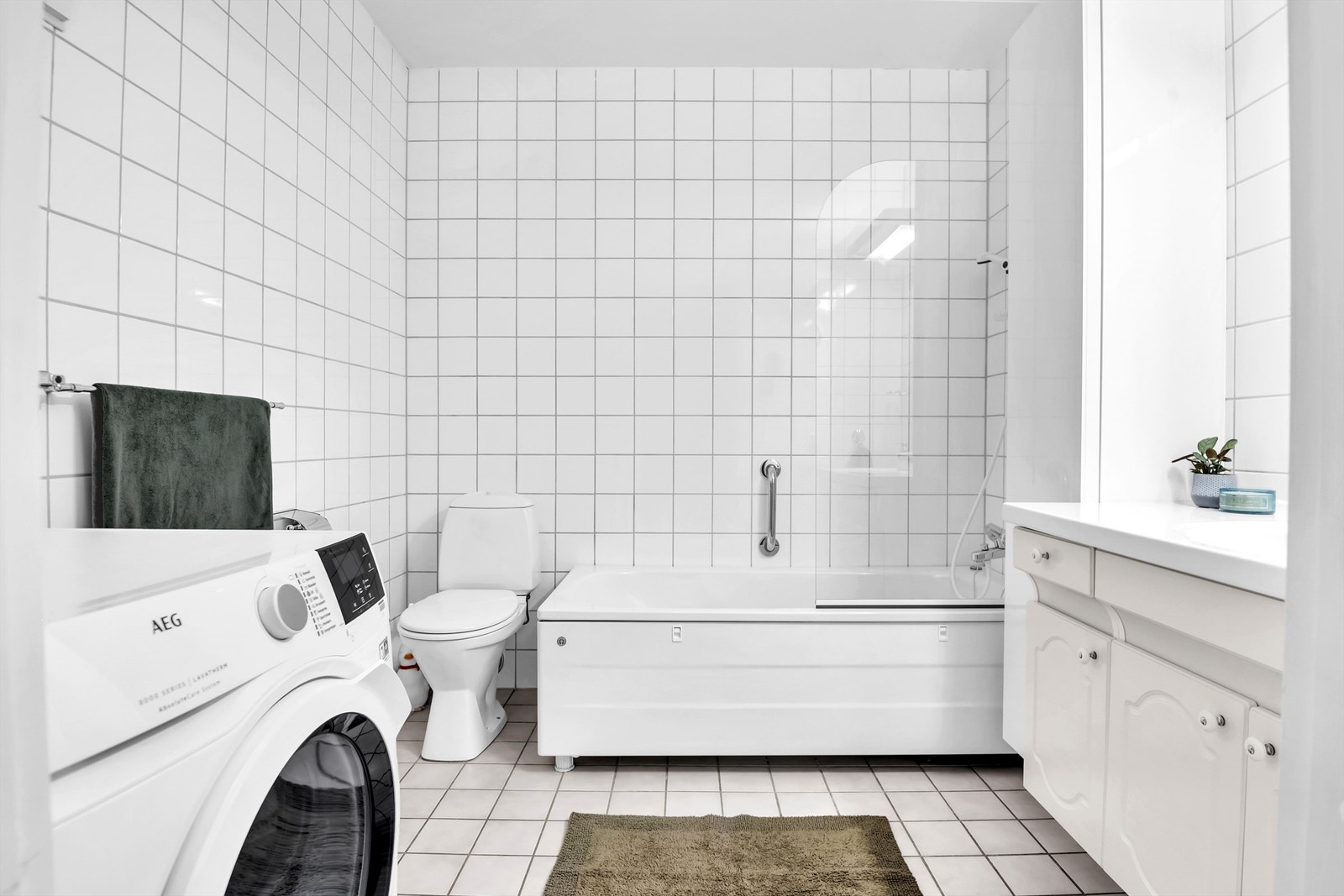 Badet er utstyrt med heldekkende servant over hvitt baderomsmøblement, speil med overbelysning, gulvmontert toalett, badekar med dusjvegg og opplegg for vaskemaskin. Galleribilde