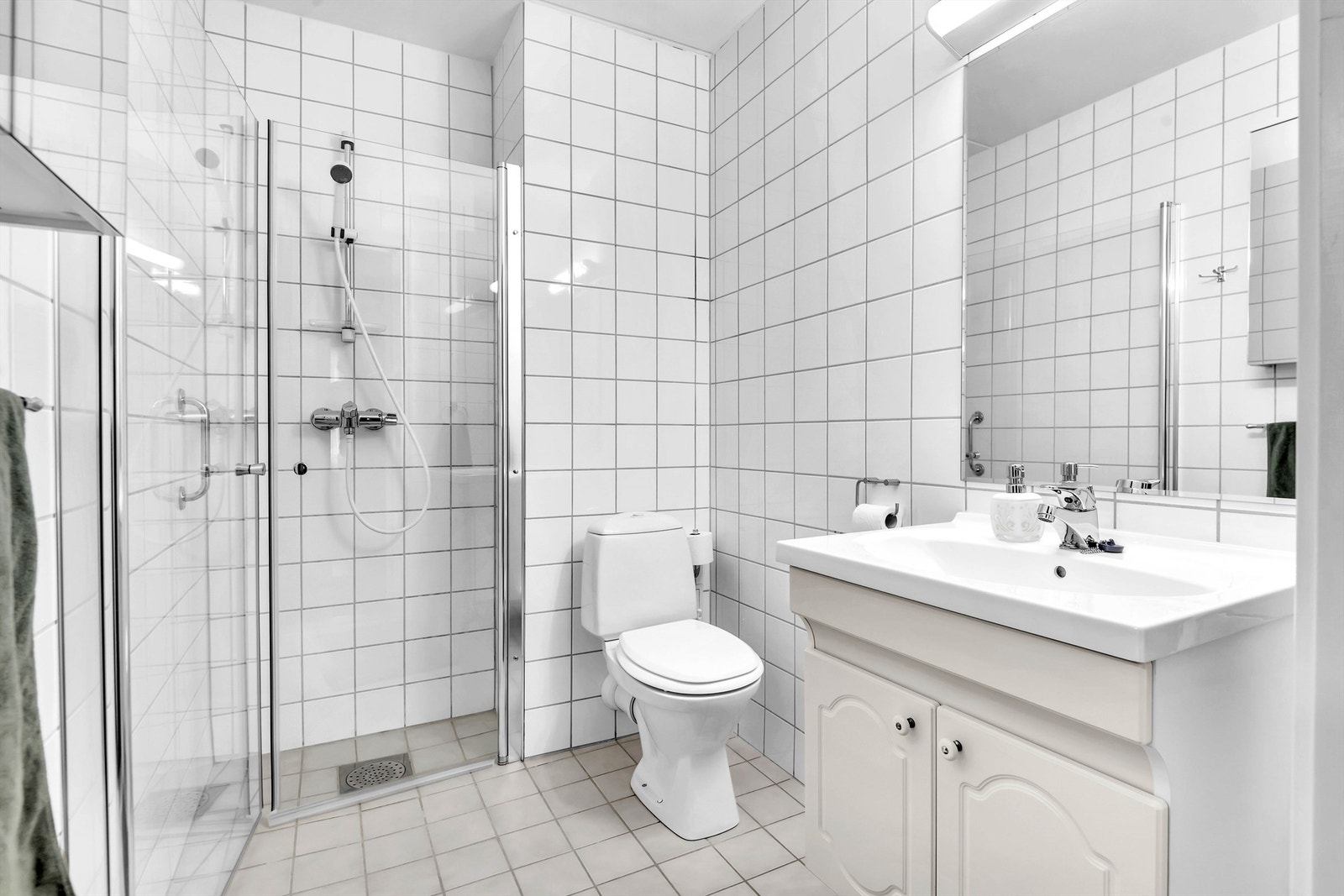 Badet er utstyrt med heldekkende servant over hvitt baderomsmøblement, speil med overbelysning, gulvmontert toalett og dusjhjørne med dusjdører. Galleribilde