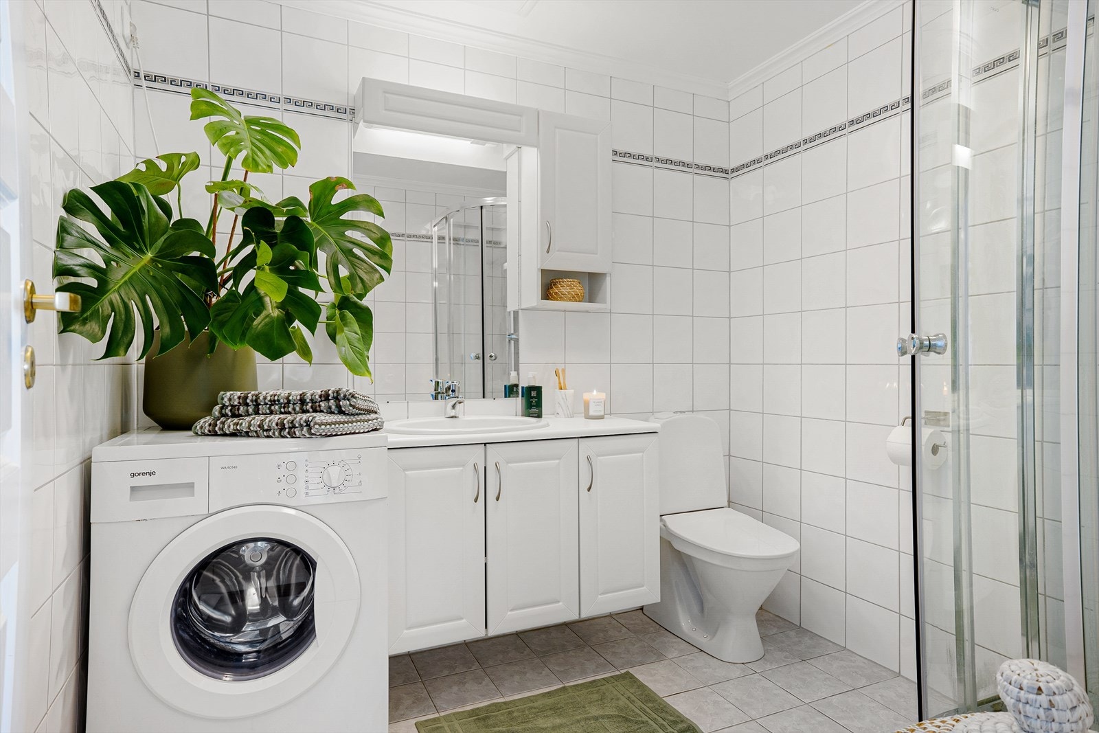 Pent, helfliset bad med lyse fliser på gulv og på vegger - badet er innredet med hvit baderomsinnredning, dusjkabinett og gulvmontert toalett Galleribilde