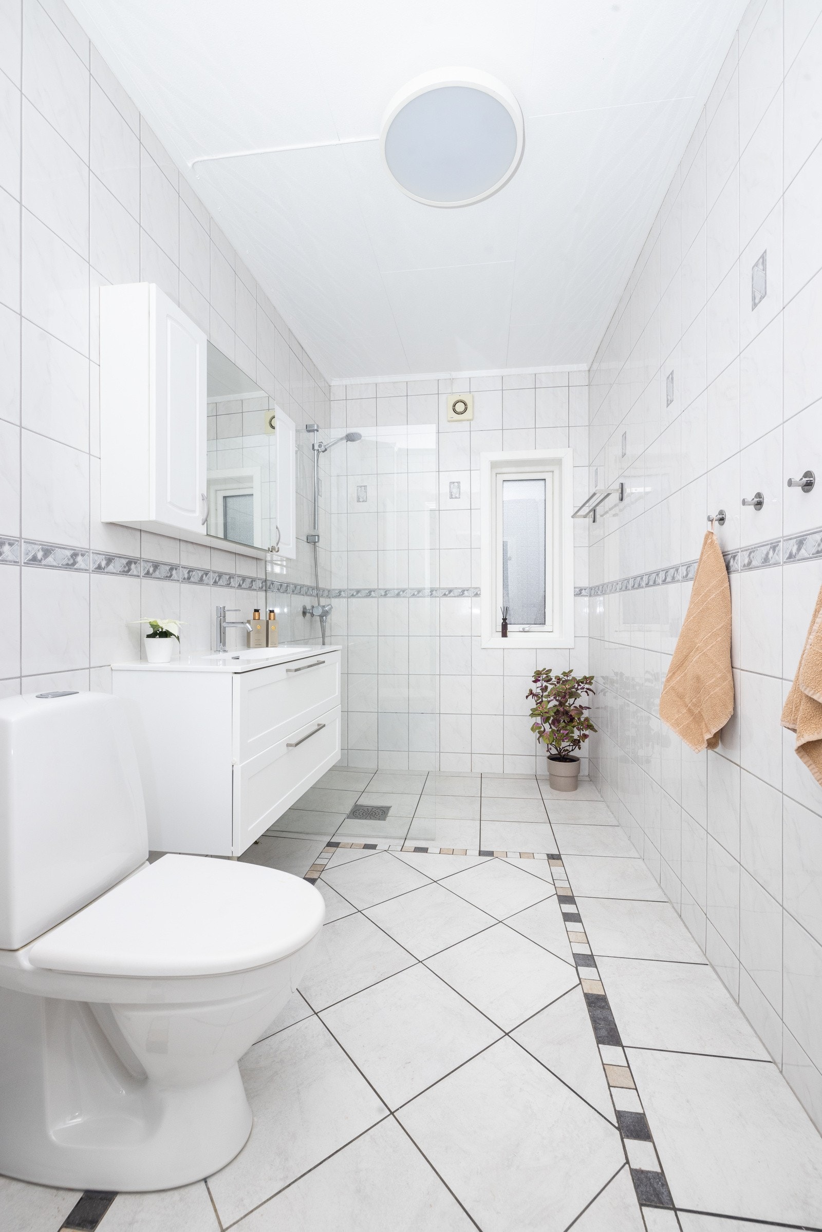 Lyst og flislagt baderom innredet med gulvstående toalett, servantinnredning, speilskap, høyskap og dusj med skjermvegg i glass. Galleribilde