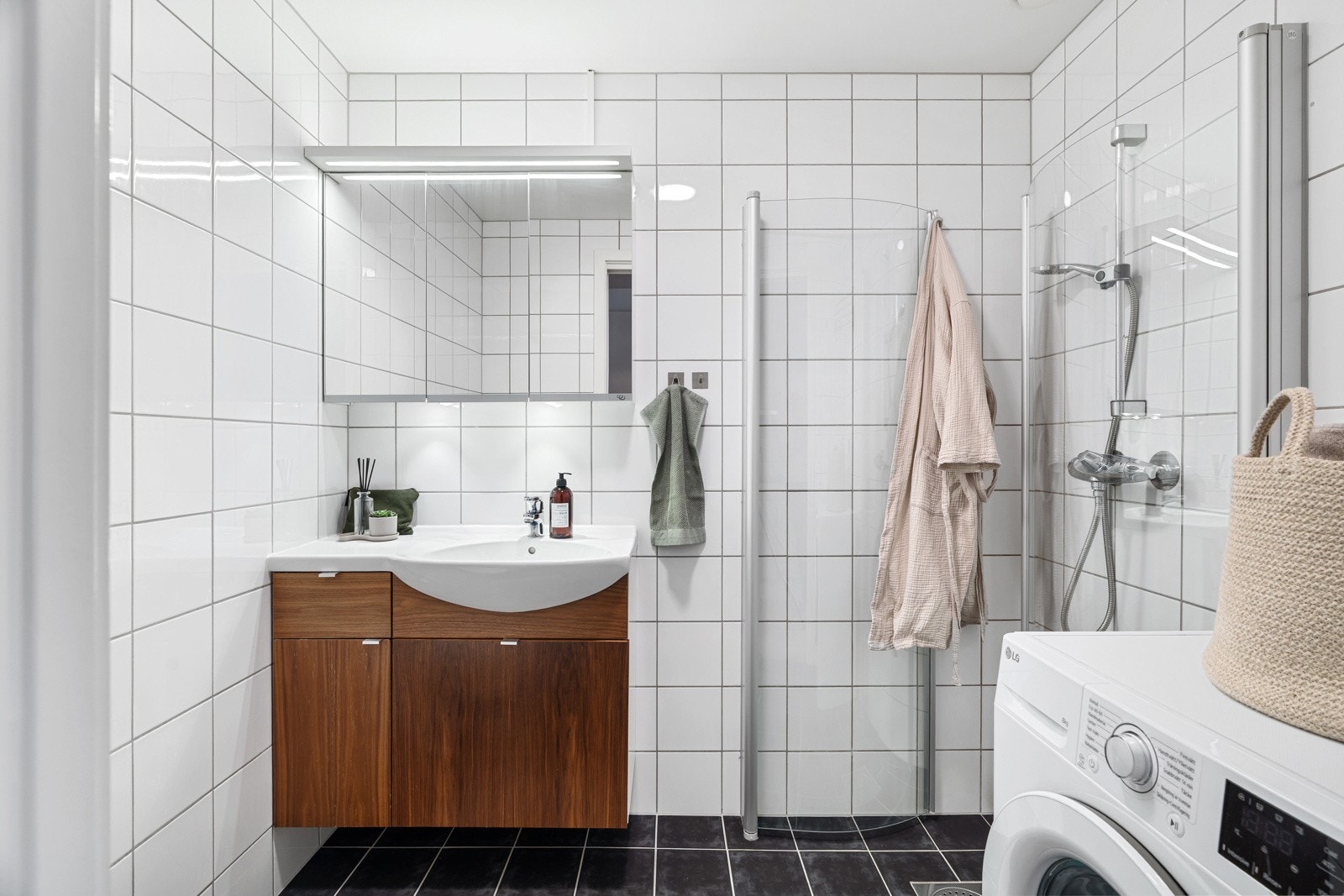 Badet er flislagt med behagelig gulvvarme. Innredningen består av skap til oppbevaring av baderomsartikler, opplegg for vaskemaskin, dusj m/innfellbare dører og toalett. Galleribilde