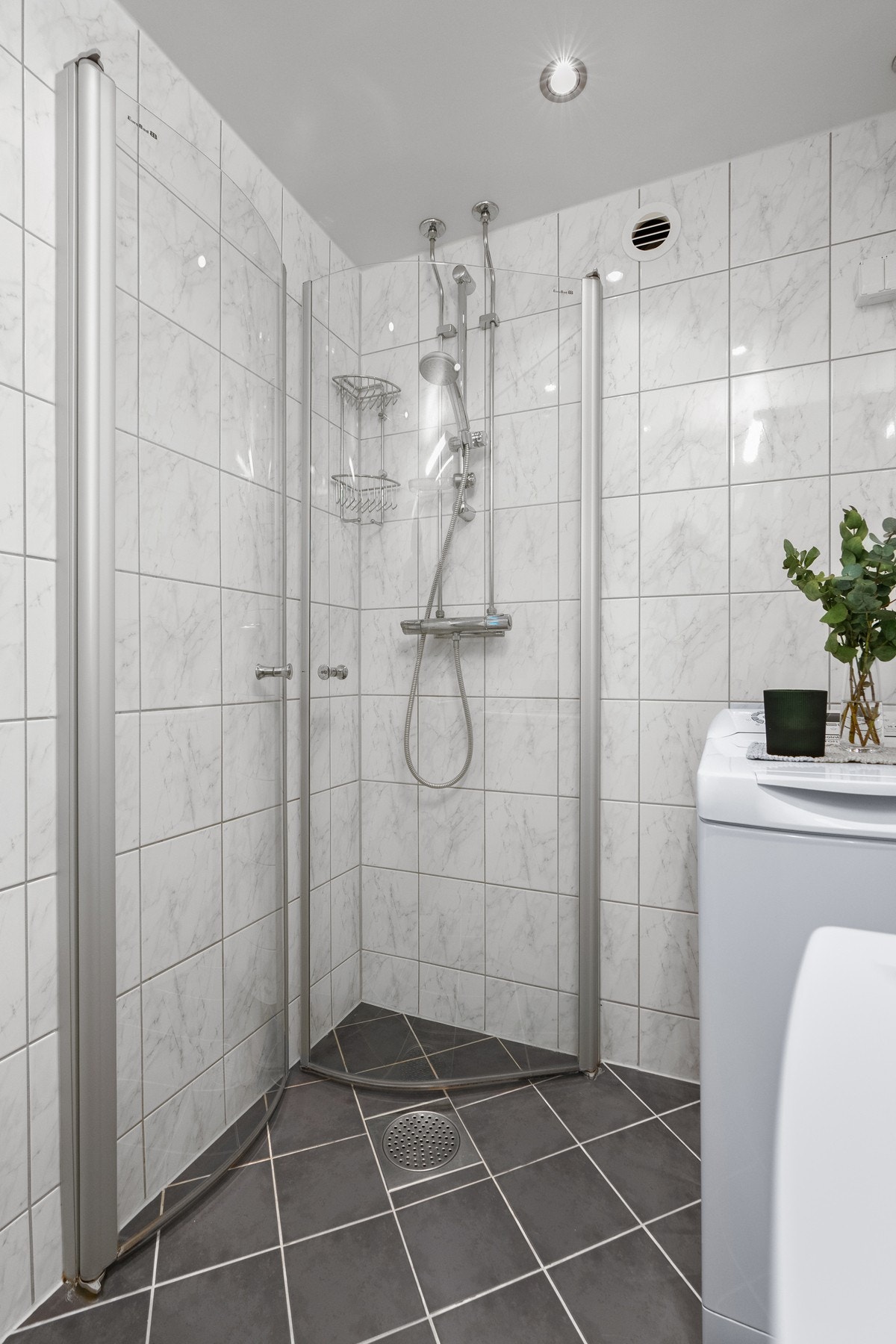 Opplegg for vaskemaskin er etablert, og badet har downlights, varmekabler og et dusjhjørne med innefellbare glassdører. Den øvrige innredningen består av et hvitt servantskap og et speilskap med belysning. Galleribilde