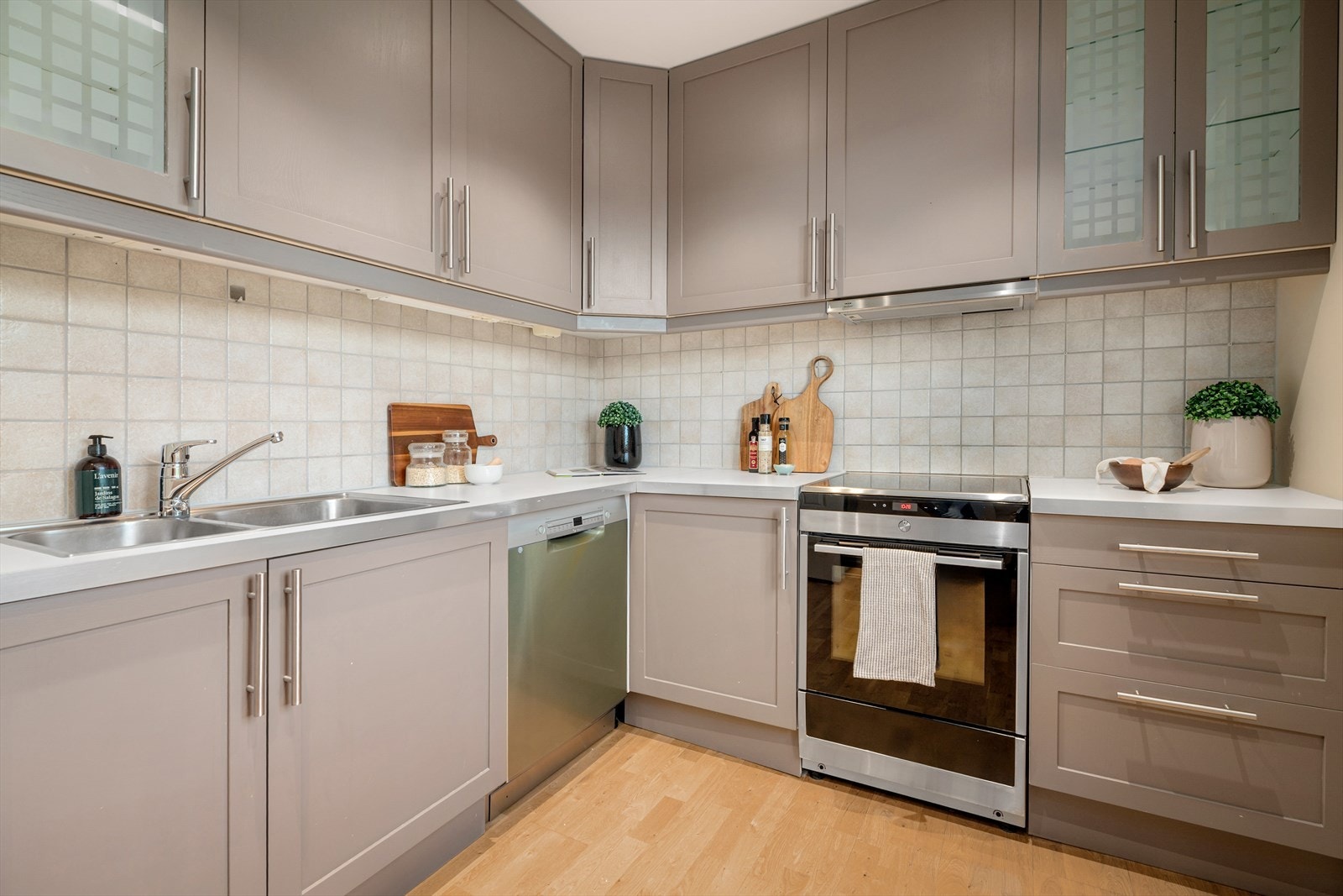 Kjøkkeninnredning med malte fronter, laminat benkeplate med nedfelt kum i rustfritt stål, fliser på vegg over benk og ventilator med kullfilter. Galleribilde