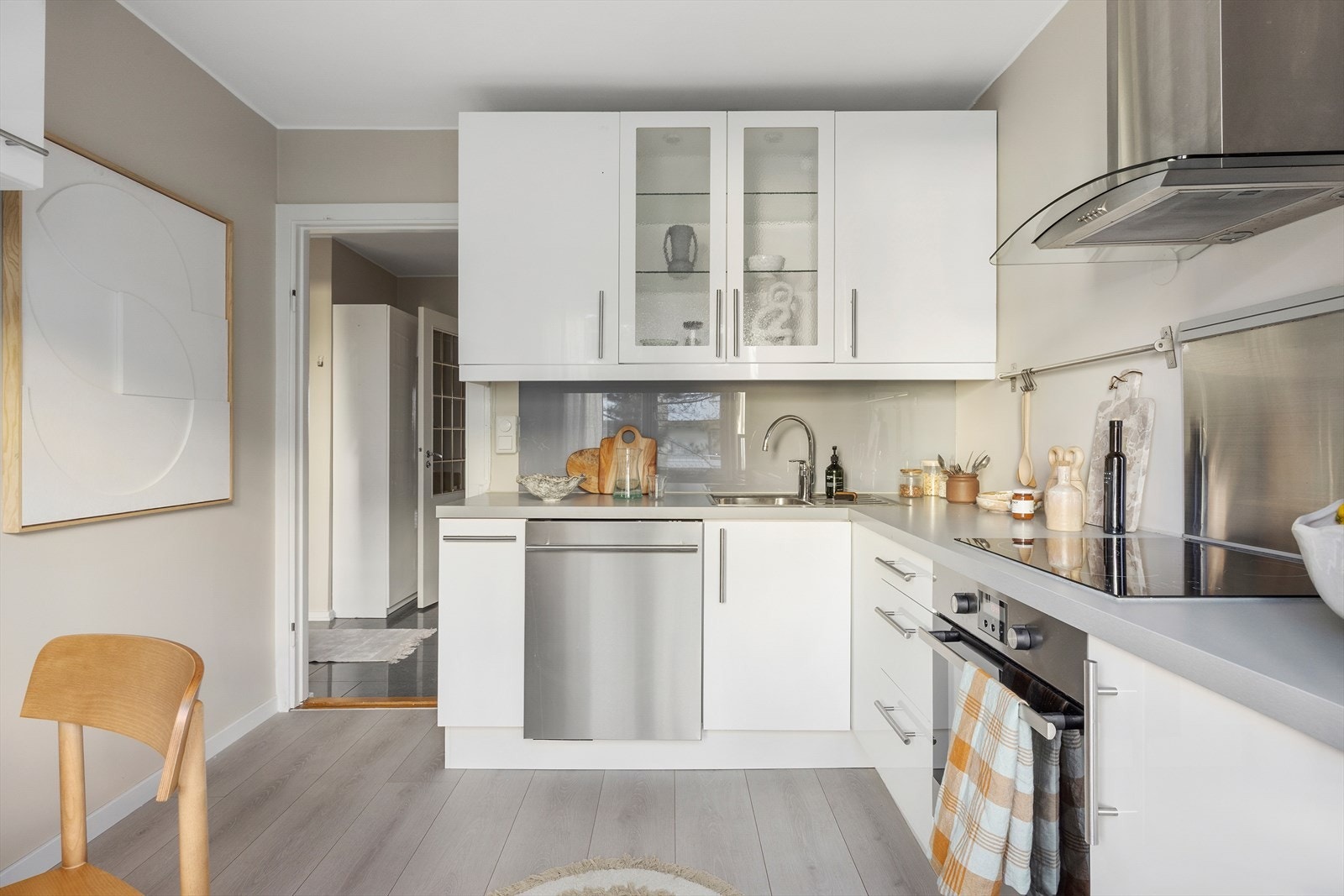 Tidløs kjøkkeninnredning med hvite glatte fronter, laminat benkeplate, integrerte hvitevarer, kullfiltervifte og frittstående kjøleskap. På gulvet er det et slitesterkt laminatgulv. Galleribilde