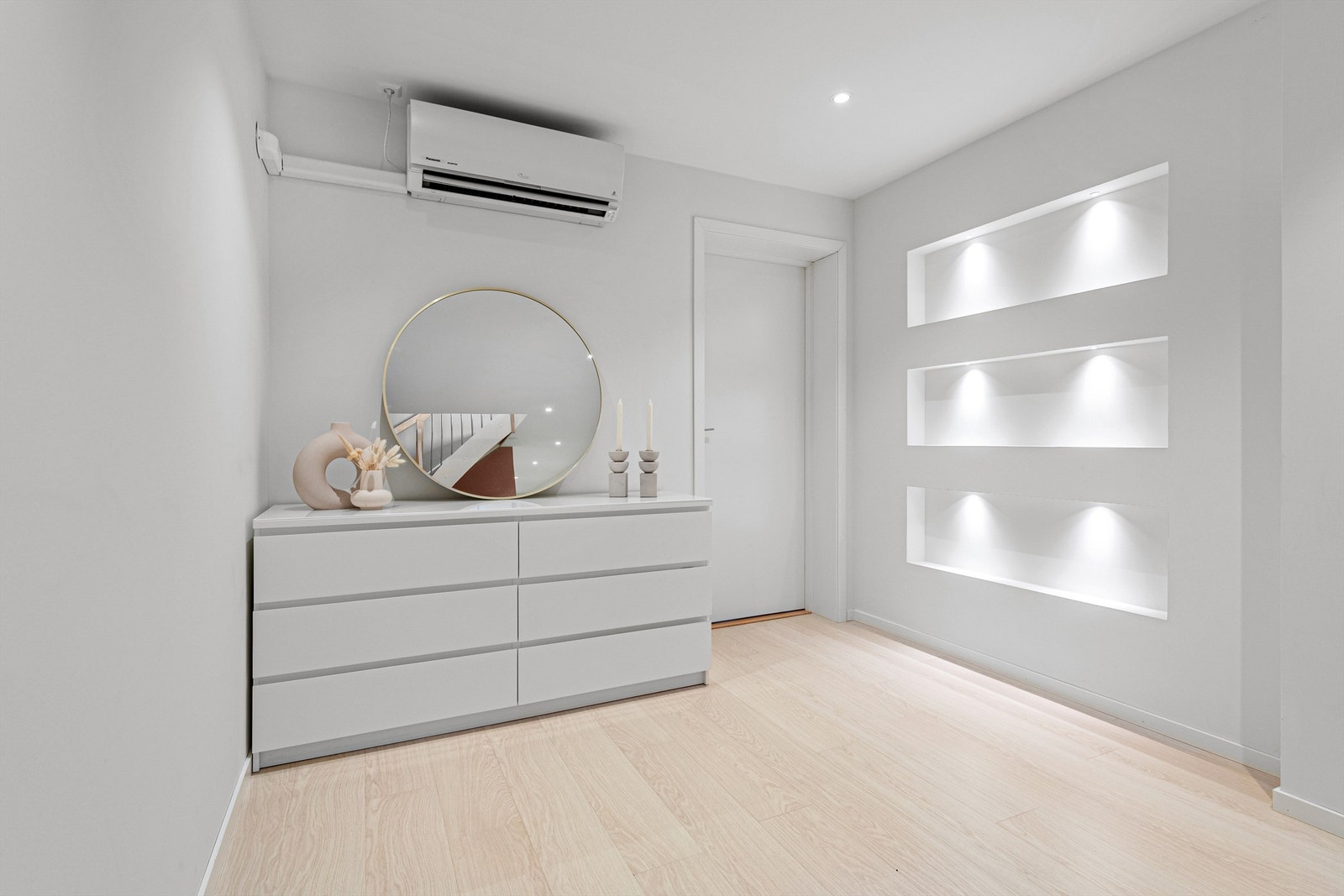 Gang i underetasjen med en-stavs laminat på gulv, malte vegger og tak med listefrie overganger og spotter i himling. Innfelte nisjer med lys i vegg. Det er montert varmepumpe i gangen. Galleribilde