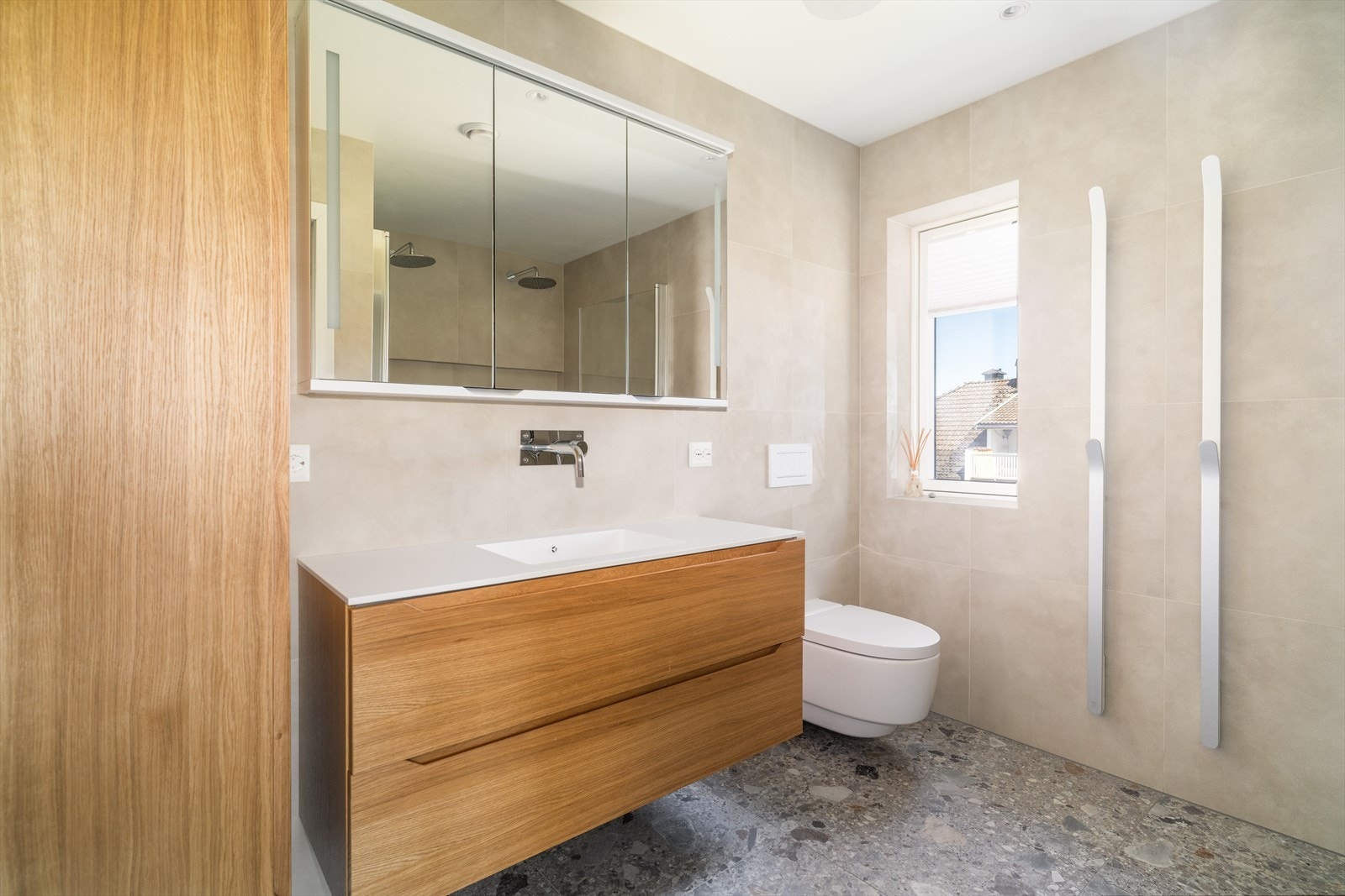 Badet er fra 2021, og har flis med varmekabler på gulv og flis på vegger. Her er det en pen innredning fra Linn Bad med heldekkende hvit vask, underskuffer med slette fronter og overskap med speildører. I tillegg et høyskap med ekstra oppbevaringsplass. Galleribilde