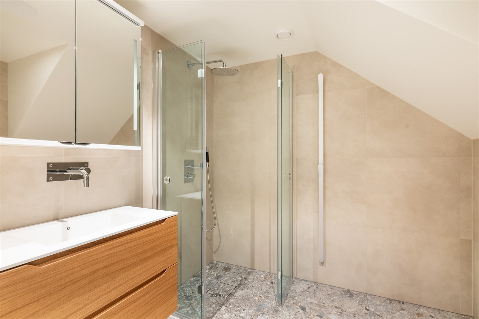 Pent bad fra 2021 med flis på gulv med varmekabler og flis på vegger. Her er det en pen innredning fra Linn Bad med heldekkende hvit vask, underskuffer med slette fronter og overskap med speildører. Galleribilde